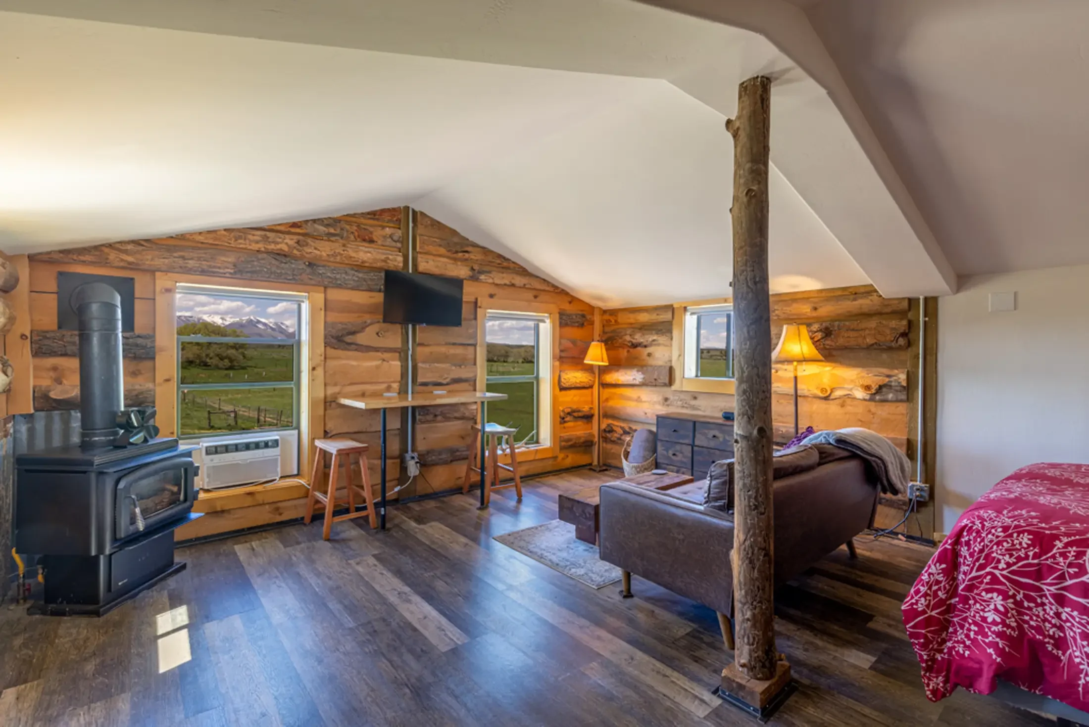 Living room with wood-paneled walls, multiple windows showing scenic mountain views, and rustic decor including a wood stove, TV, and two lamps, with a log column separating the space.