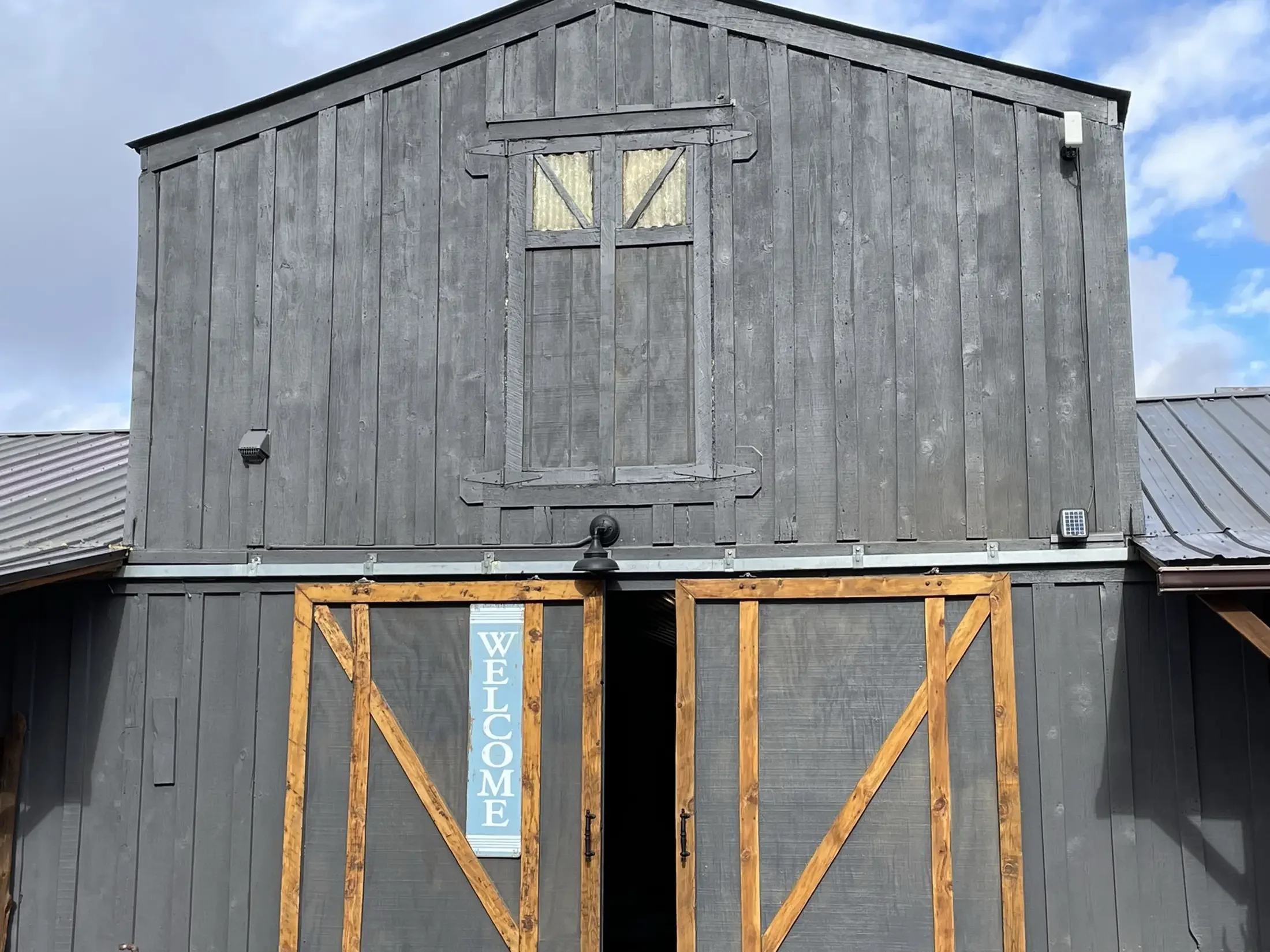 A gray wooden barn with a small window covered in weathered yellow fabric, a black lantern light, and a blue vertical sign that says WELCOME inside a wooden frame.