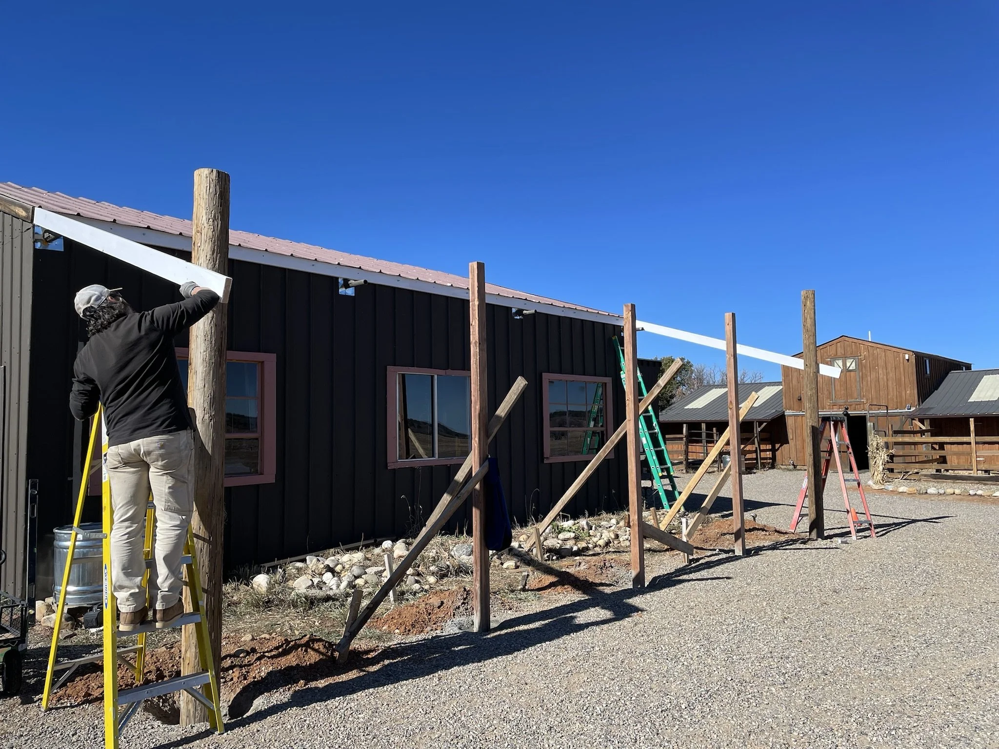 Person standing on a yellow ladder working on a wooden utility pole outside near a black building with pink window frames under a clear blue sky.