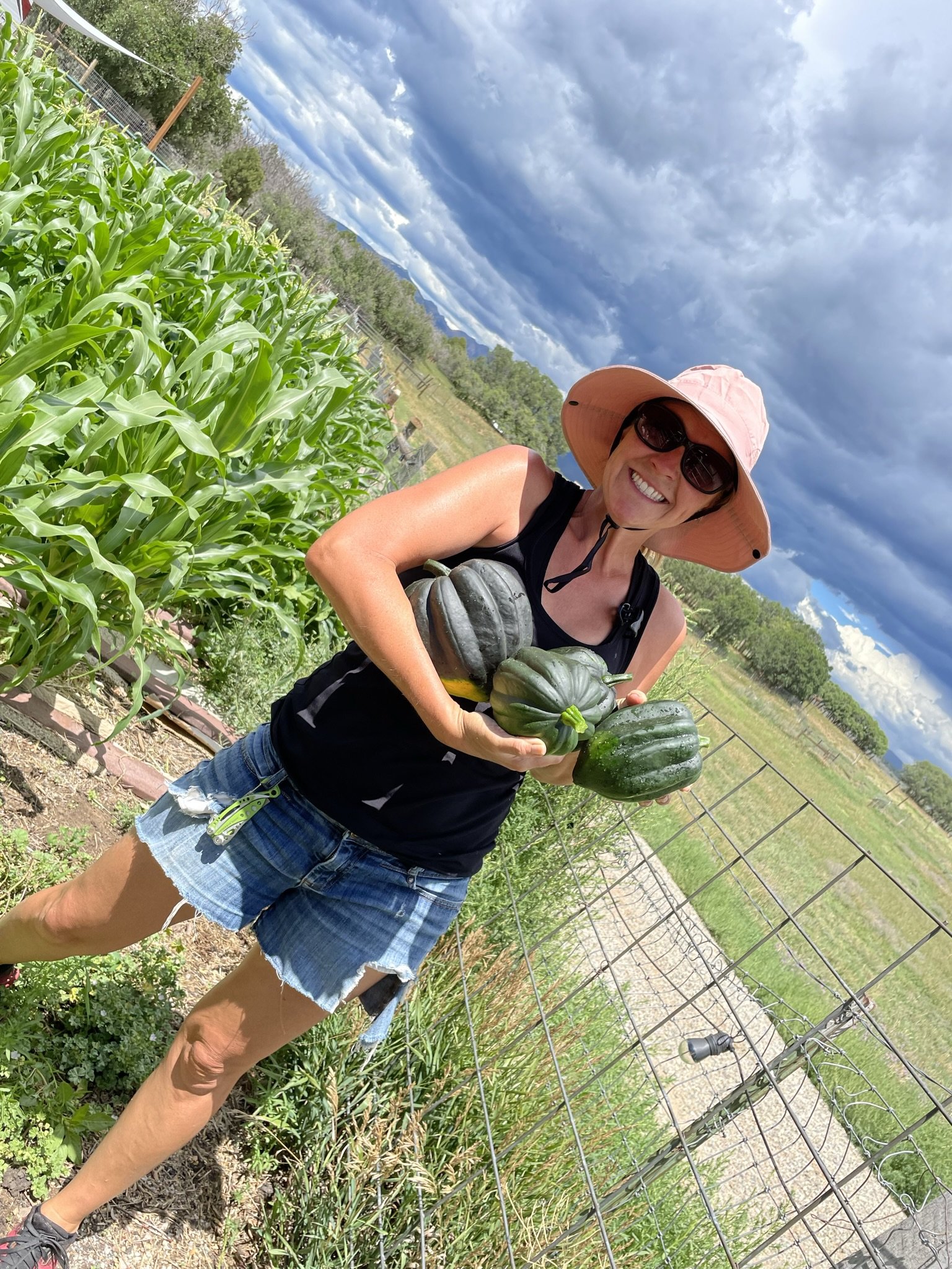 A smiling woman with a pink sun hat and black sunglasses holding green and striped gourds in a garden with green plants and a wire fence, under a partly cloudy sky.
