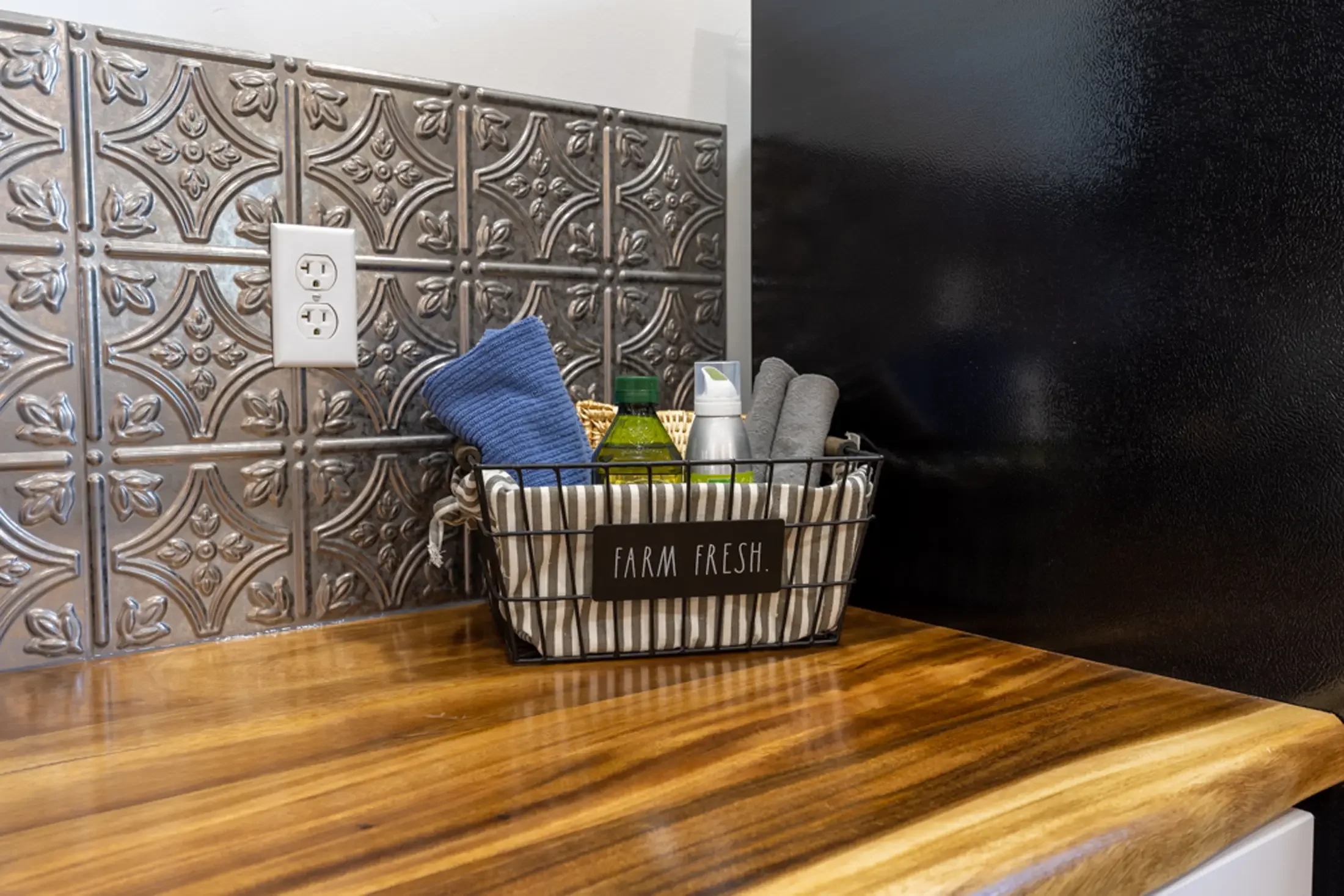 Decorative silver-colored wall with intricate floral pattern, electrical outlet, basket containing cleaning cloth, spray bottle, and a green bottle, with a sign reading 'FARM FRESH' in a striped container on a wooden surface.
