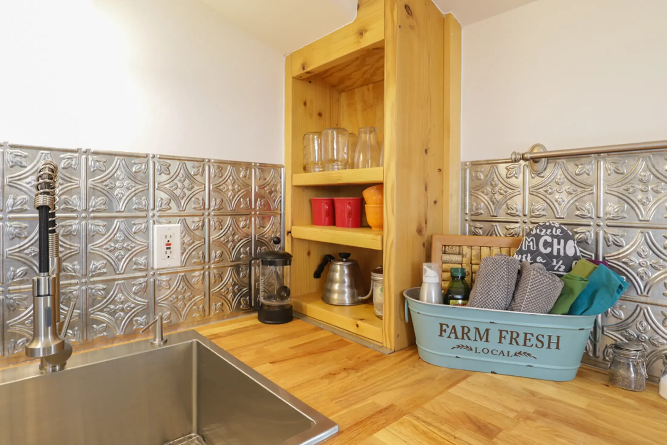 Kitchen with a stainless steel sink, wooden countertop, and open wooden shelves with glasses, red cups, and orange bowls. A decorative metal backsplash is behind the countertop, and a blue farm fresh basket with cloth and other items is on the counte