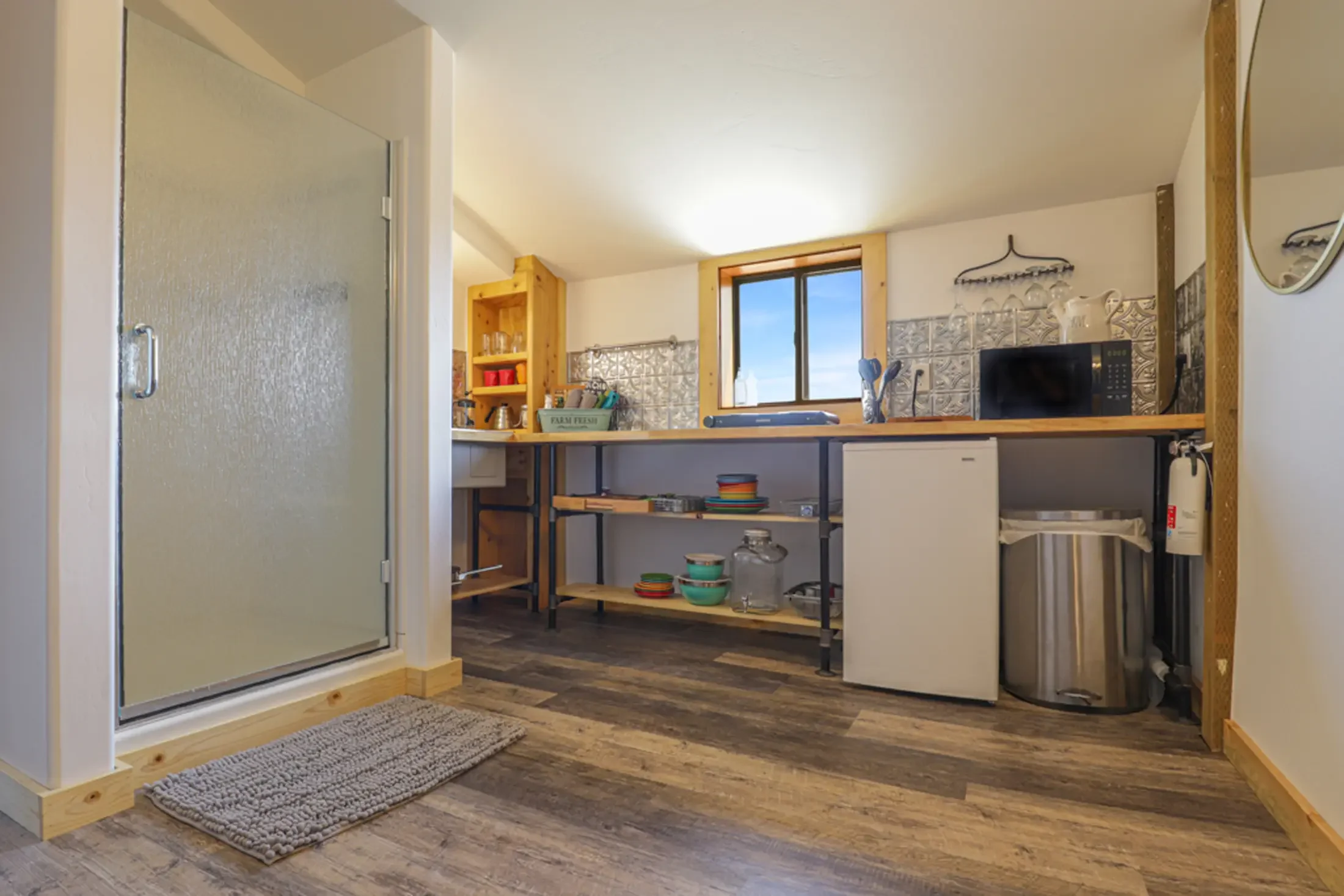 A small kitchen with a wooden counter, a microwave, metal shelves with dishes, and a window showing blue sky outside. The floor has wood-like planks and there is a small rug near the entrance.