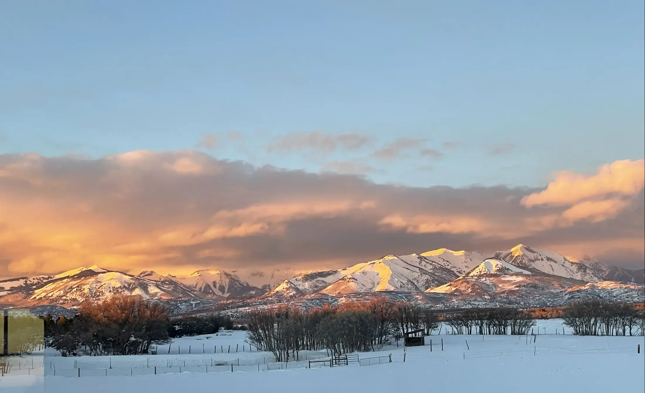 Snow-covered fields with a fence in the foreground, barren trees, and snow-capped mountains in the distance under a sky with pink and orange clouds during sunset.