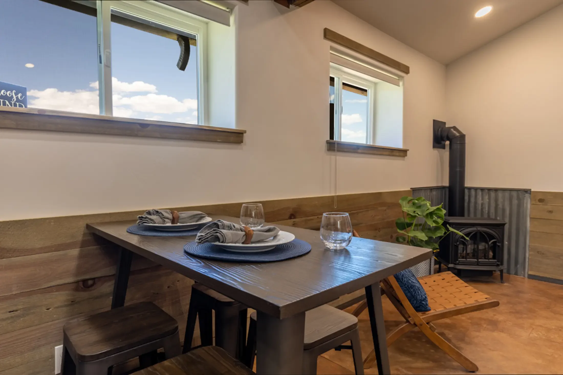 A dining area with a wooden table set for three, featuring plates, glasses, and cloth napkins, with a window showing a blue sky with clouds. There is a wood stove and a potted plant in the corner.