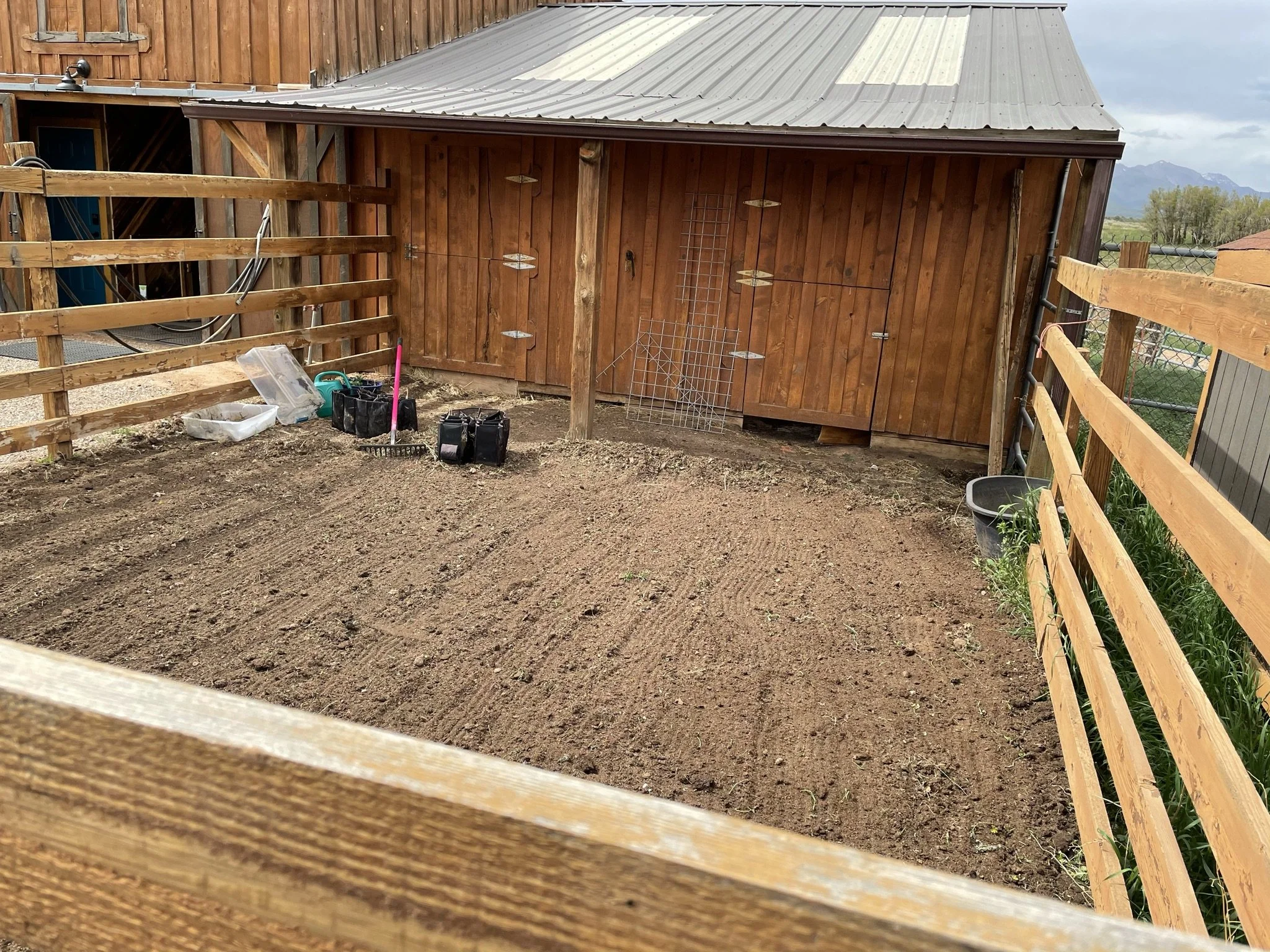 A wooden barn with a metal roof, surrounded by a dirt area and a wooden fence. Construction tools and supplies are on the ground near the barn.