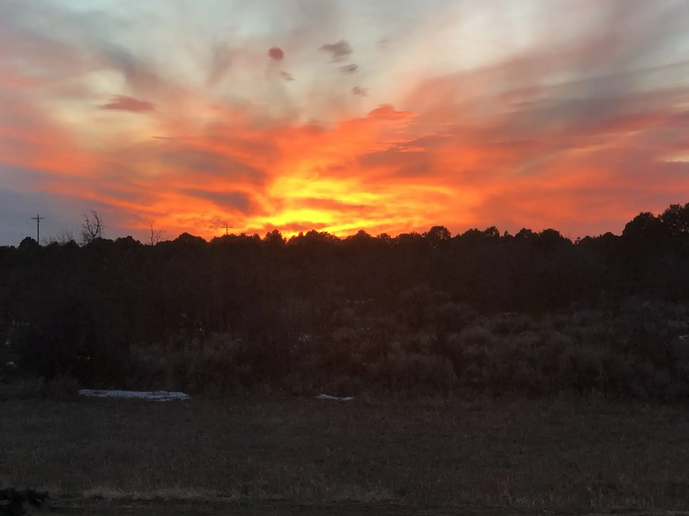 A colorful sunset with vibrant orange, yellow, and pink hues in the sky, silhouetted trees, and power lines in the foreground.