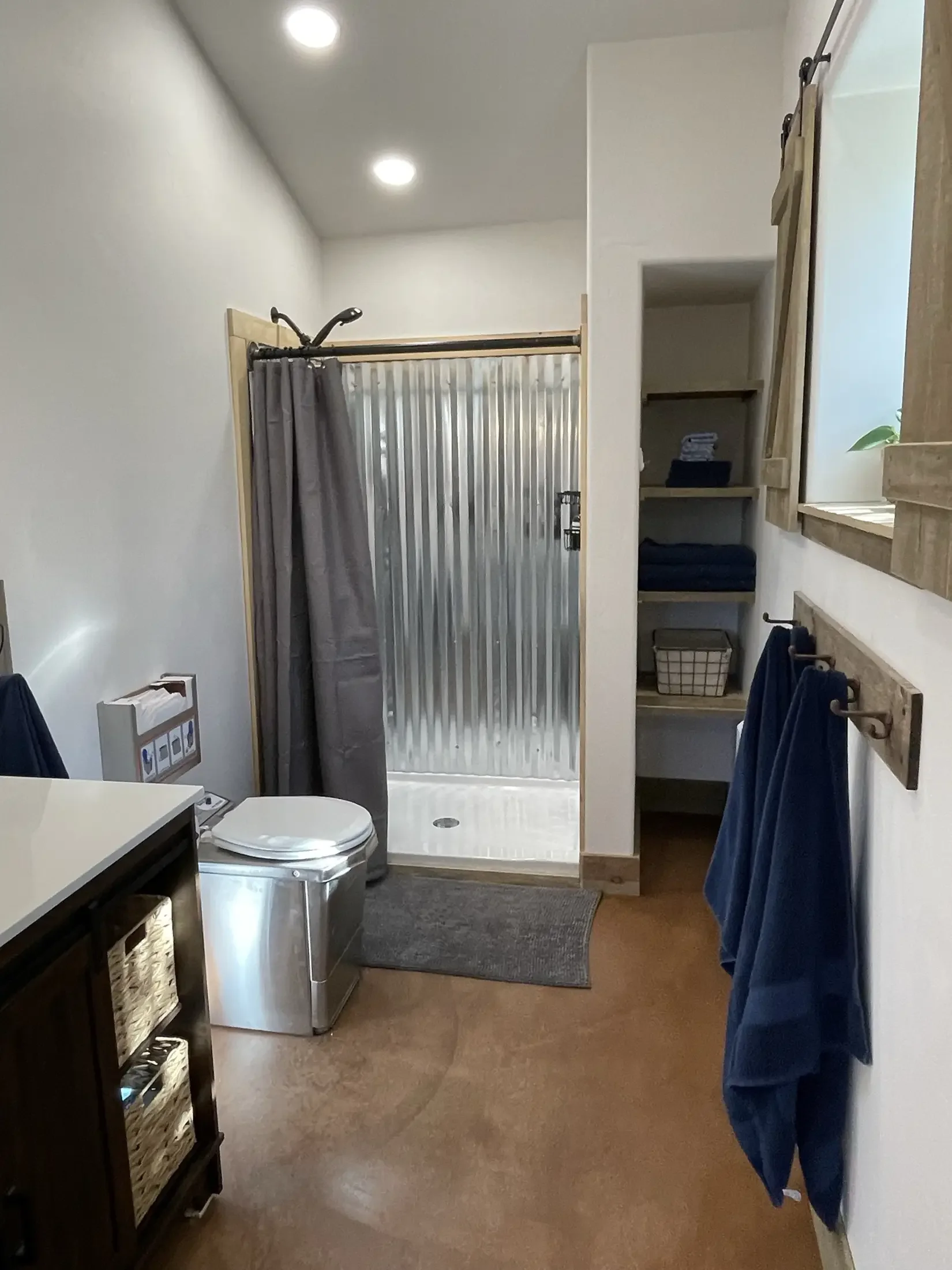 A bathroom with a shower area with a corrugated metal door, gray curtain, and circular drain on the shower floor. There are recessed ceiling lights, a closet with shelves containing folded towels and a basket, and multiple blue towels hanging on hook
