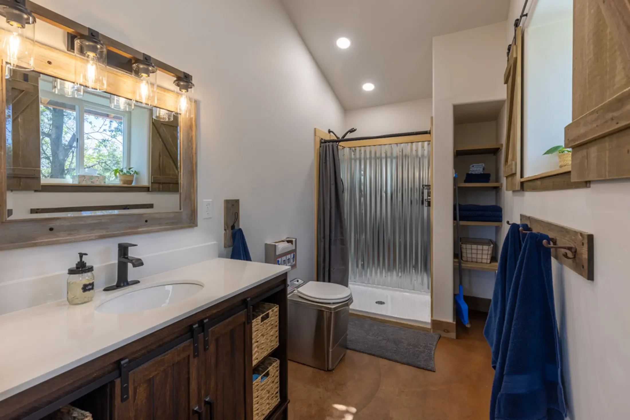 A bathroom with a white countertop sink, a dark wooden vanity, a mirror with a wooden frame, a window with shutters, a plant on the windowsill, a laundry area with shelves and towels, a metal shower with a curtain, and blue towels hanging on a rack.