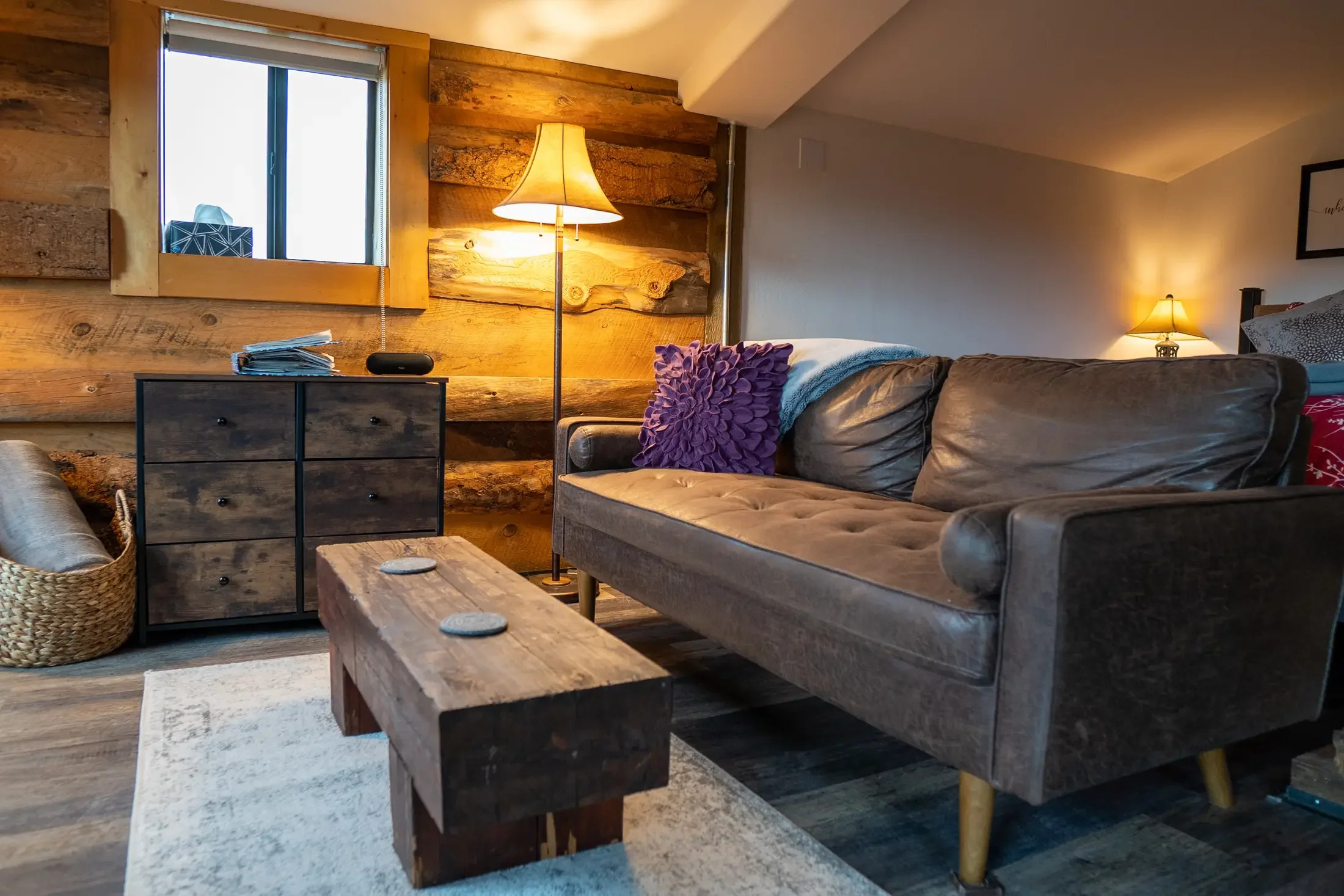 A cozy living room with a leather sofa, purple pillow, and rustic wooden furniture. There are two table lamps, a window with wooden trim, wooden wall paneling, and a small black speaker on a dark wooden cabinet.