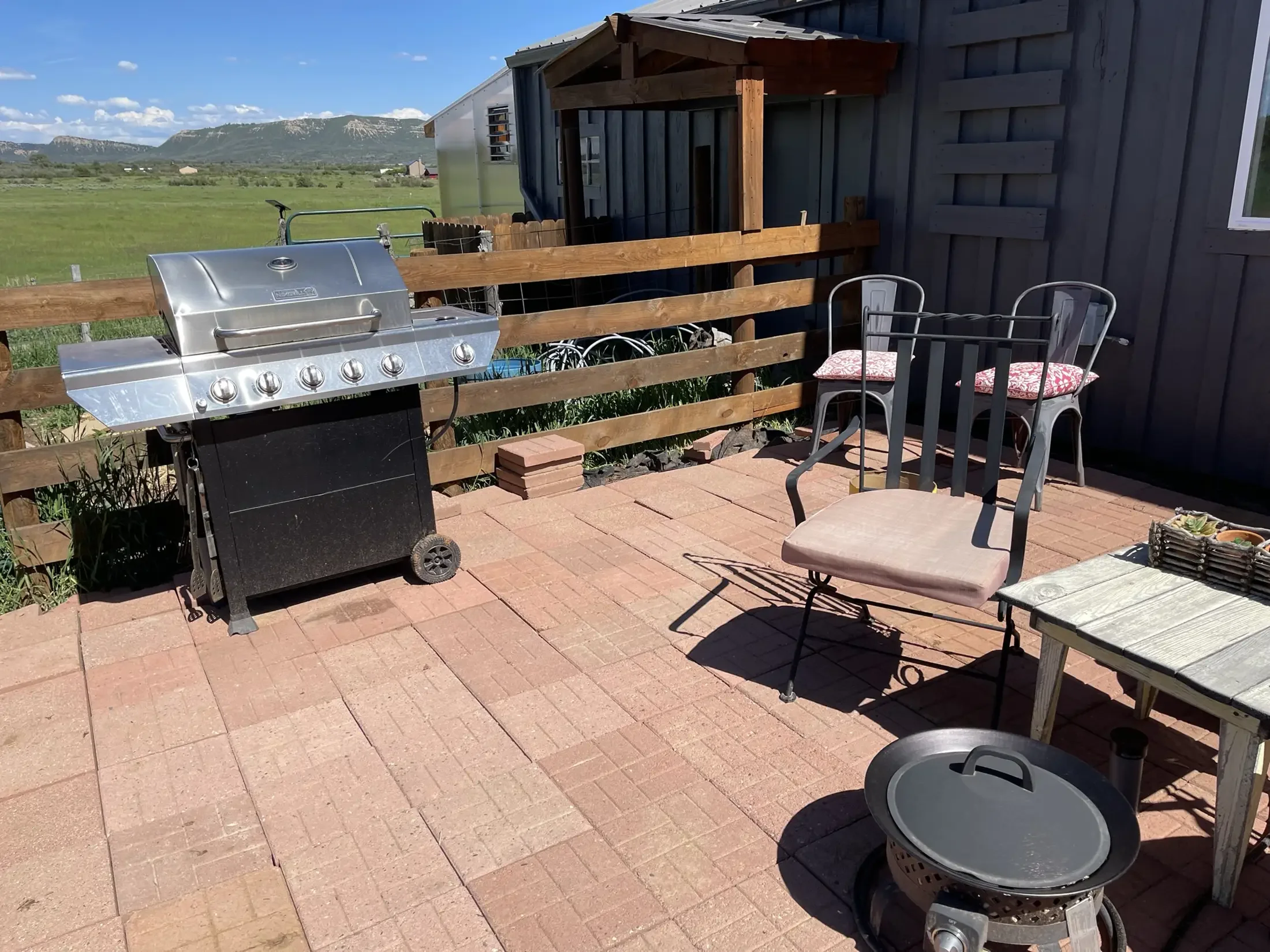 A backyard patio with a black grill, patio furniture including chairs with red patterned cushions, and a wooden fence. In the background, there are green fields, distant mountains, and a blue sky with some clouds.
