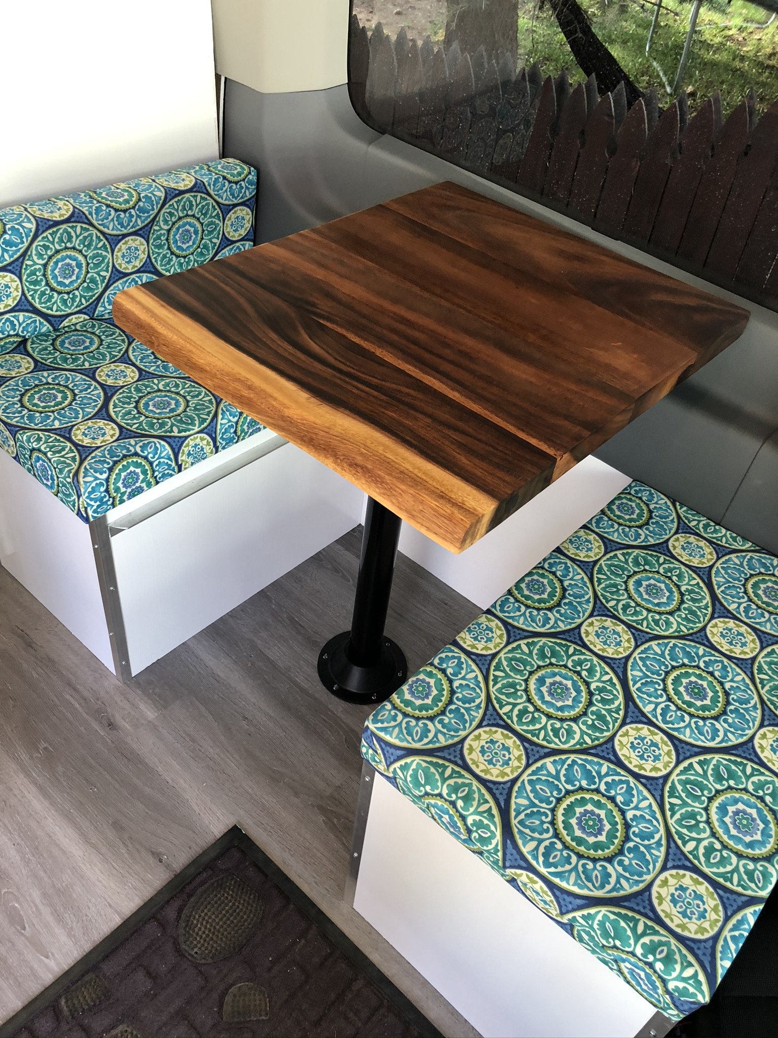 A small table with a wooden top, situated between two colorful patterned chairs with blue, green, and yellow designs, in a cozy indoor space by a window overlooking a garden.