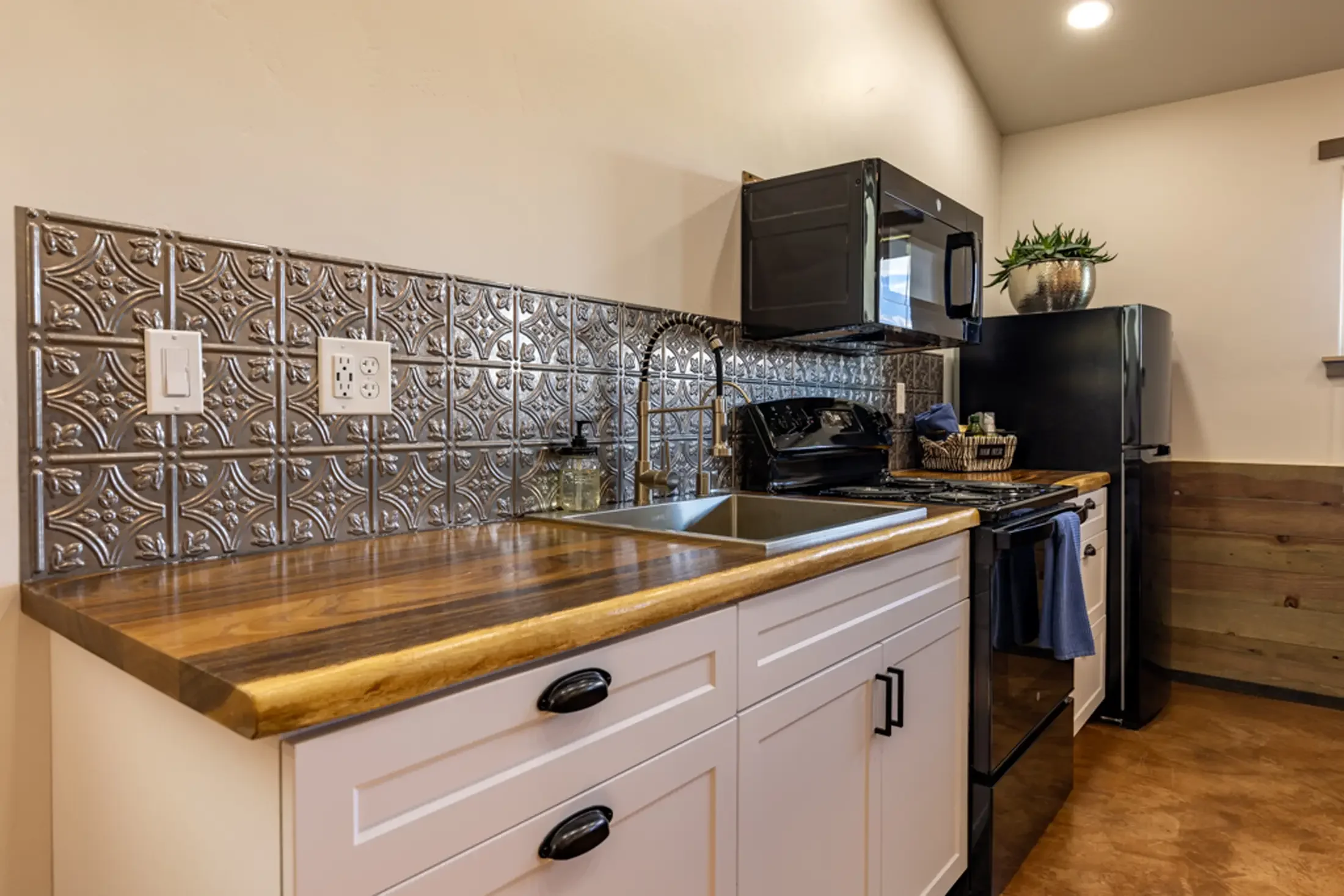 A kitchen with a wood countertop, patterned metal backsplash, white cabinets, a black microwave, black stove, gray refrigerator, and a potted plant on top of the refrigerator.