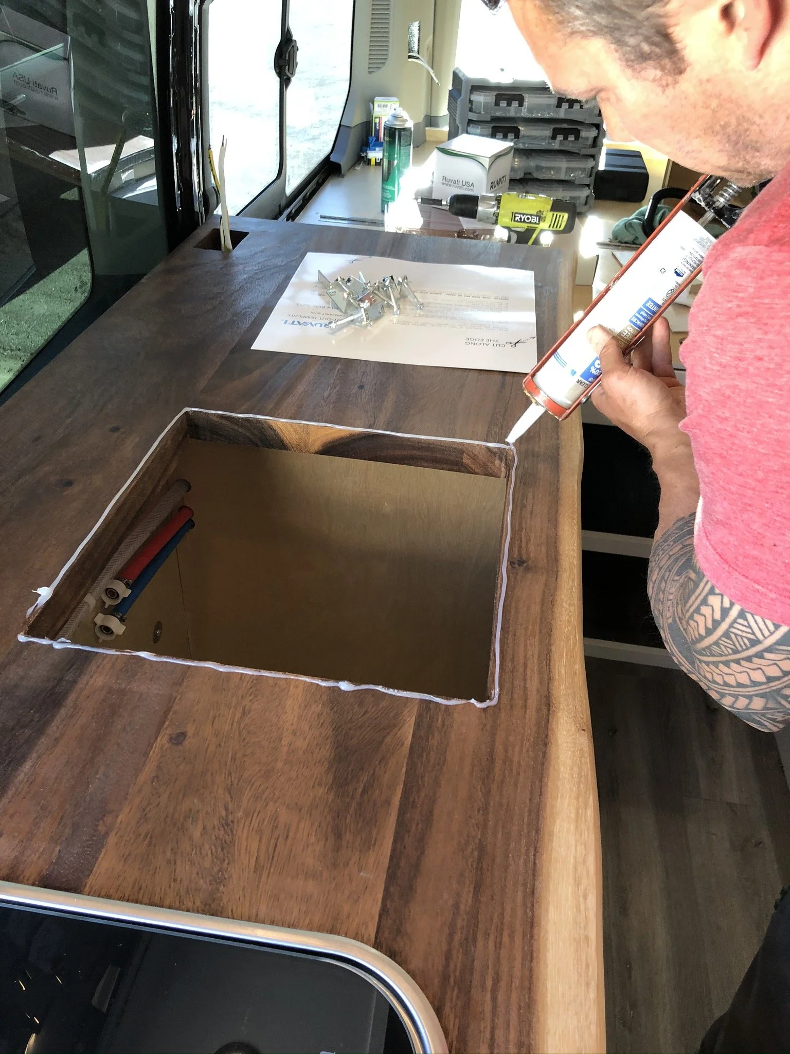 A man working at a woodworking project on a wooden counter, applying adhesive glue inside a square cutout with visible pipes inside, using a caulking gun, in a workshop environment with tools, a drill, and organized storage in the background.