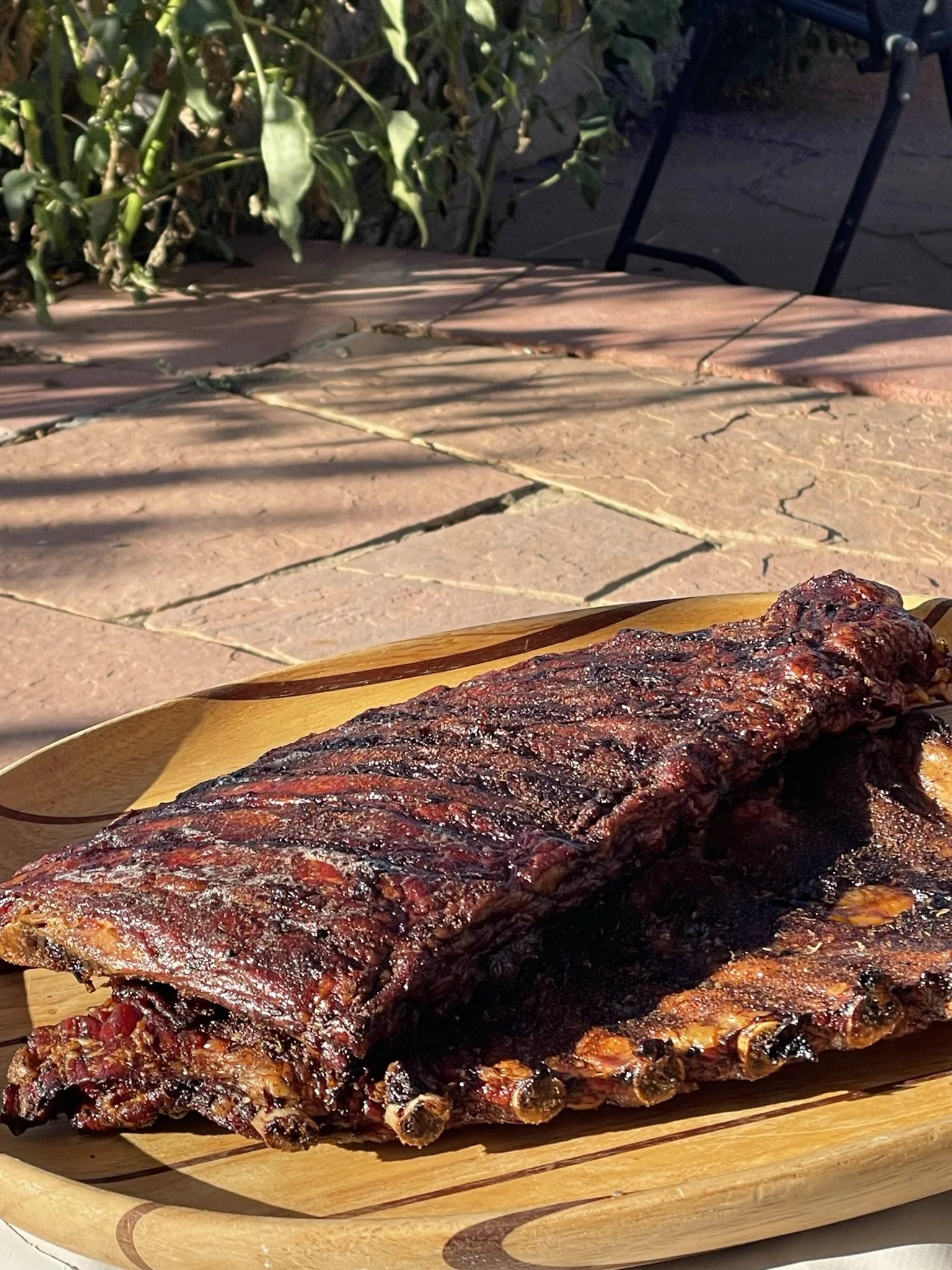 Close-up of a smoked beef brisket on a wooden cutting board with a background of outdoor patio furniture and greenery.