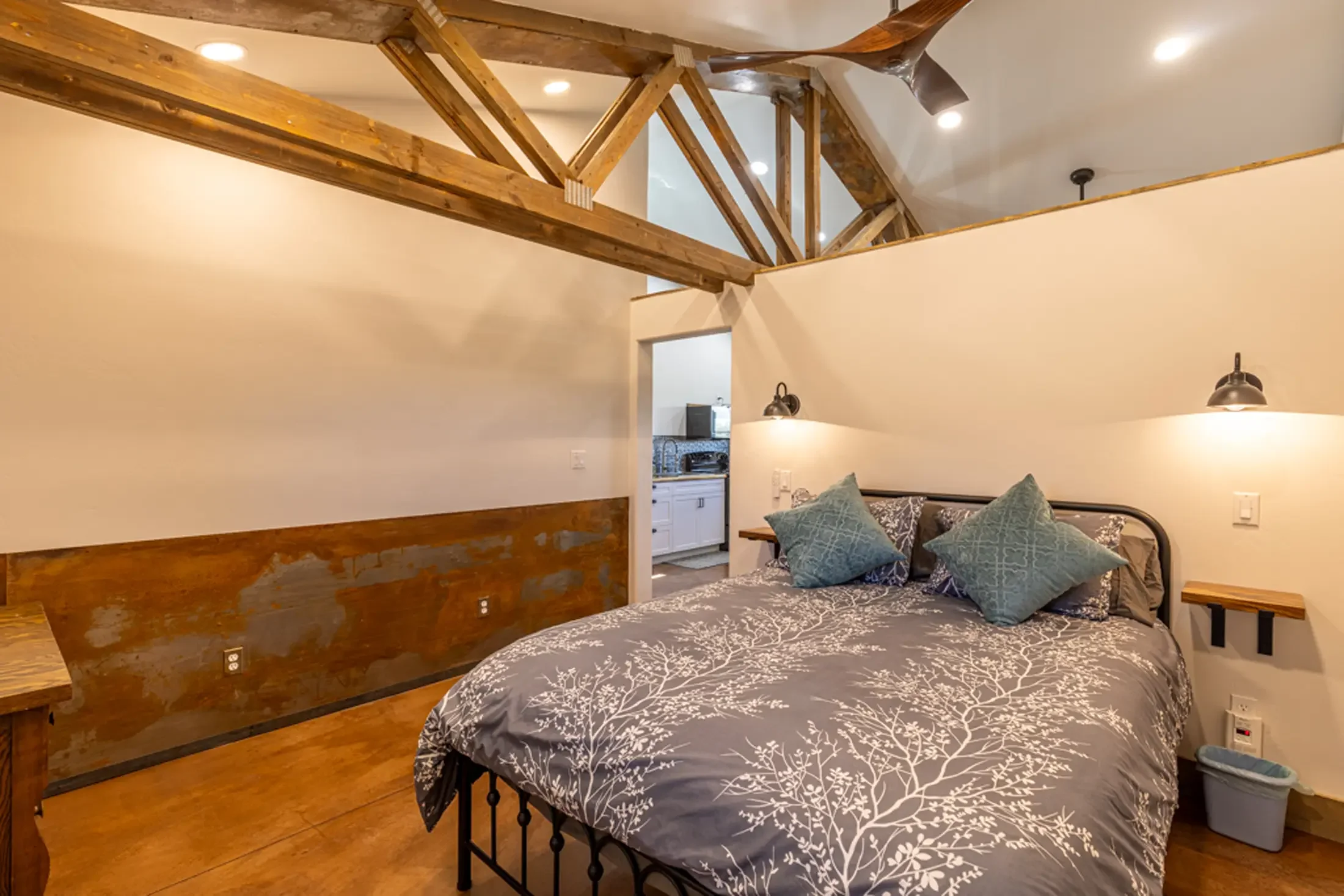 A bedroom with a bed covered in a black and white bedspread with tree branch patterns, several blue pillows, and a wooden headboard. The room features wooden beams on the ceiling, white walls, and is located adjacent to a kitchen visible through a do