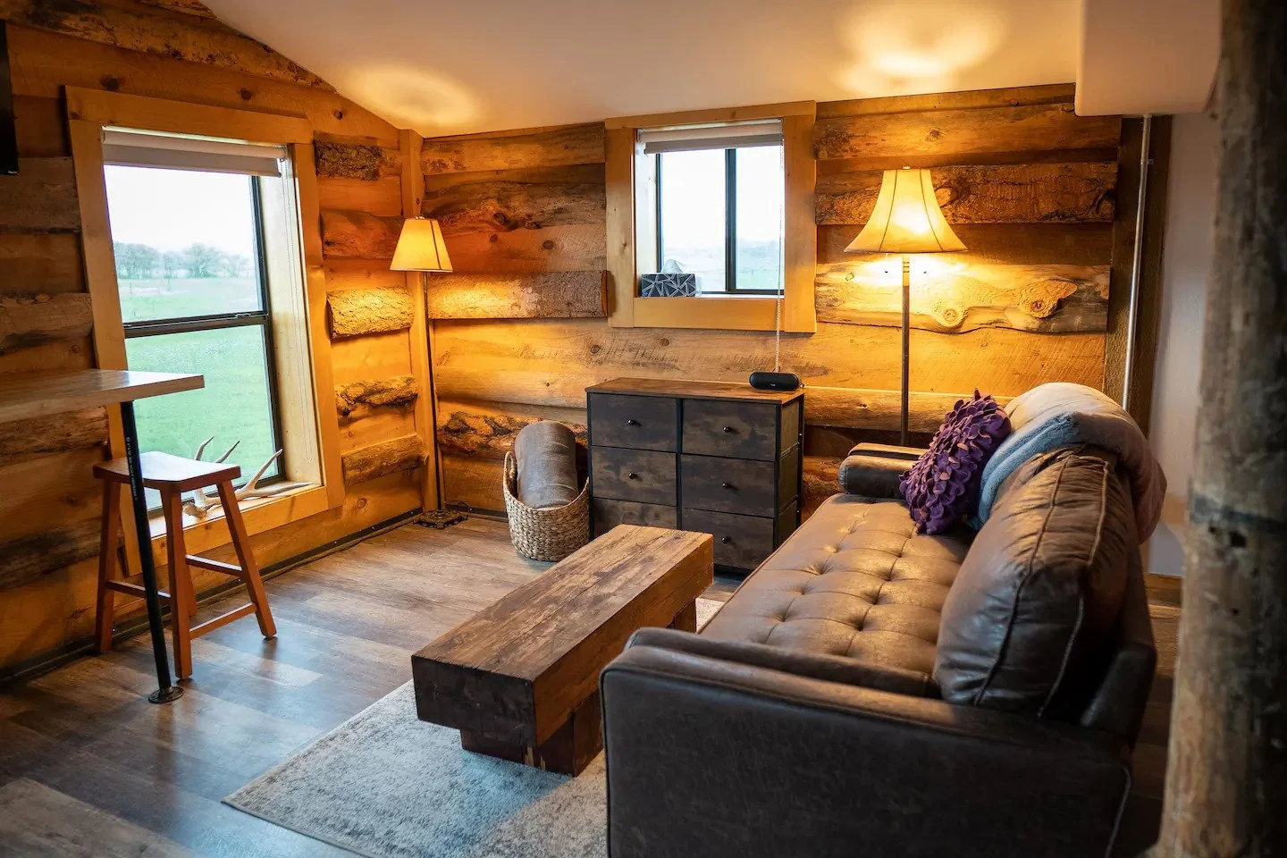 Cozy rustic living room with wooden plank walls, two windows, a leather sofa with purple and blue pillows, two floor lamps, a dark wood dresser, a woven basket, a wooden coffee table, a small table with a chair near one window, and a ceiling light.