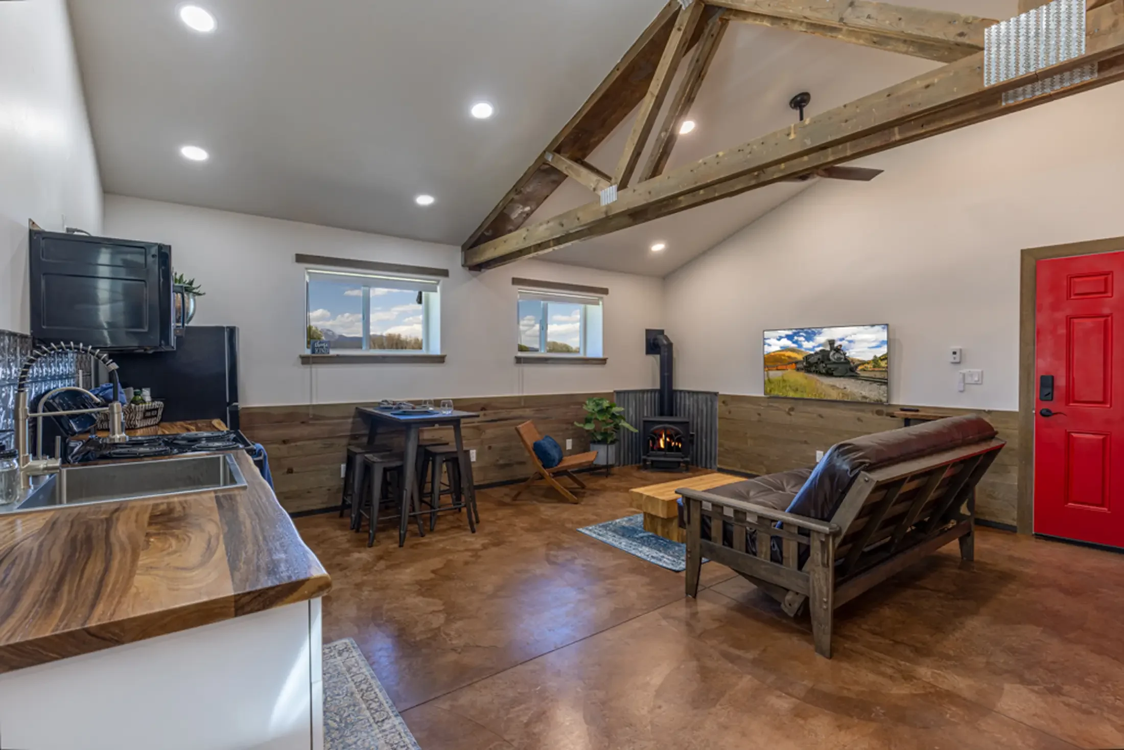 Open-plan living room with wood accents, white walls, and a vaulted ceiling with exposed wooden beams. Features include a wood stove, two windows showing a blue sky with clouds, a flat-screen TV, and a red door. There is a wooden dining table, chairs