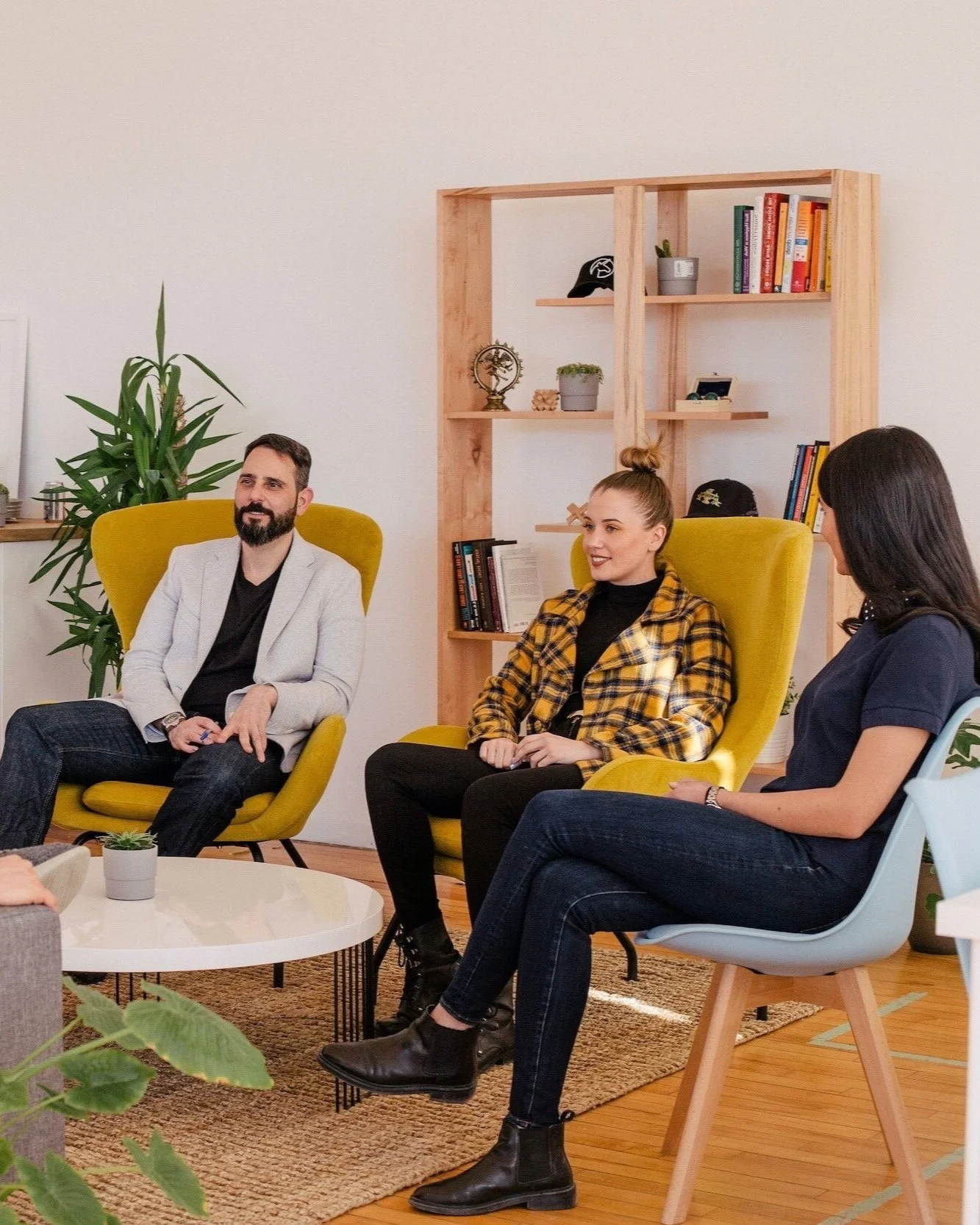 A woman sitting on a pebble beach with her head resting on her arms and her knees pulled to her chest, looking down. The sky is cloudy and the sea is in the background.