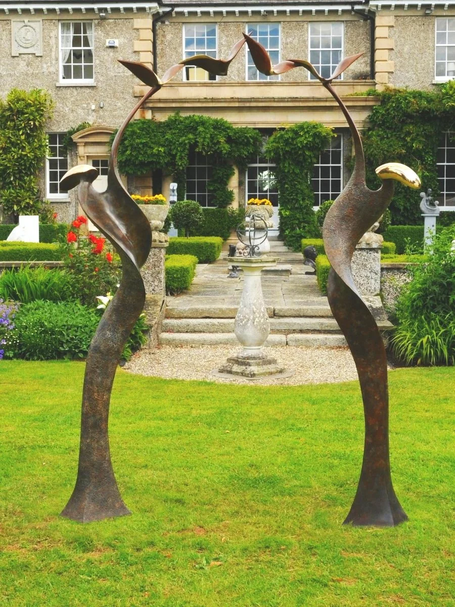 arch-garden-sculpture-cast-bronze-figure-spiral-aloft-birds-curved-ribbon-shapes-unfurl-golden-heads-arch-support-world-patinated-deep-browns-tree-boughs-peaceful-mythological-forest-sandra-bell-metal-modern-art-sustainable-process-materials-ireland