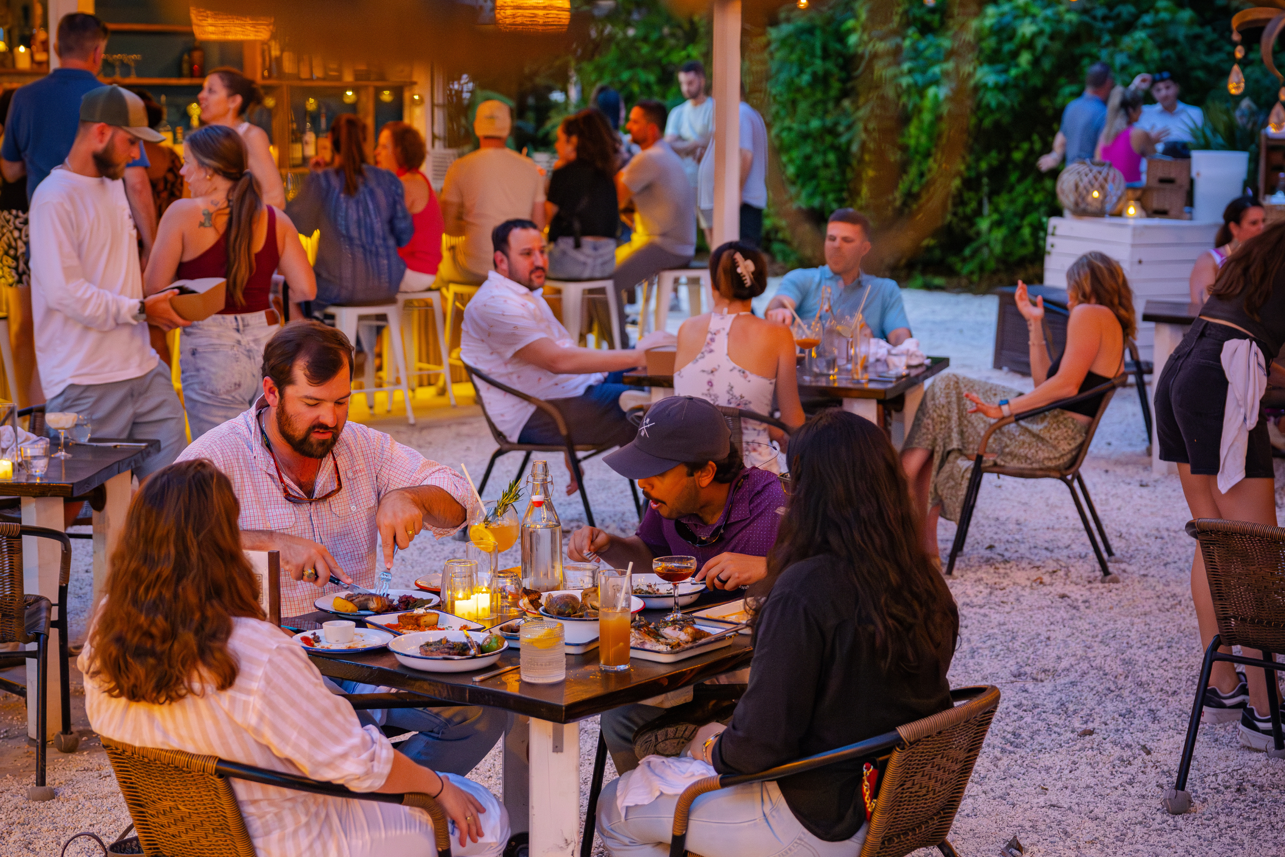 People dining outdoors at a lively restaurant during evening, with food, drinks, and socializing.