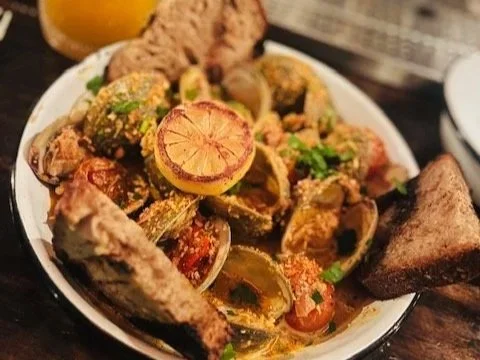 A bowl of cooked seafood, possibly clams or mussels, garnished with lemon slices and green herbs, with pieces of bread on the side.