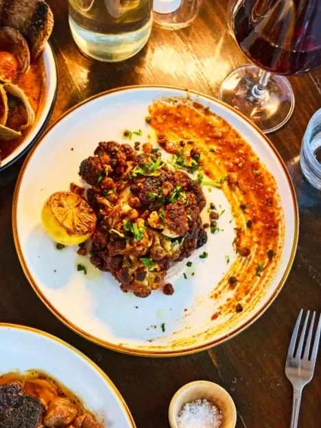 Plate with cooked ground meat topped with chopped herbs, a roasted lemon half, and a smear of sauce or puree. Surrounding items include a glass of red wine, a glass of water, and other dishes on a wooden table.