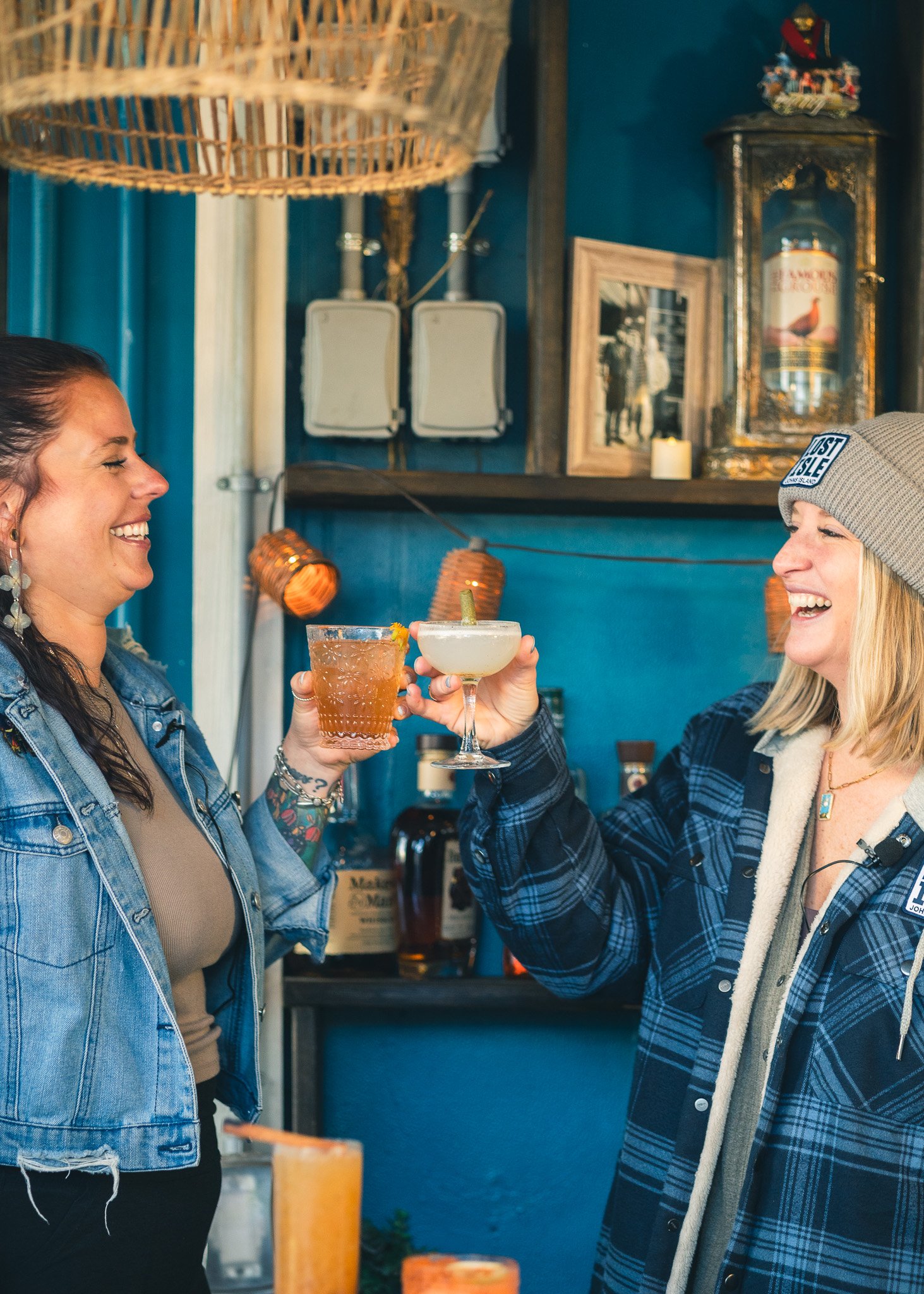 Two women smiling and toasting drinks in a cozy bar with blue walls and vintage decor.