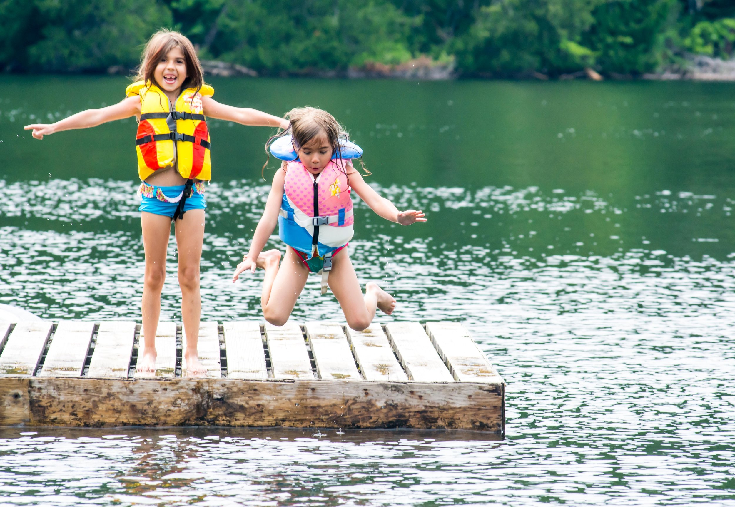 Two children jumping off a wooden dock into a lake, wearing colorful life jackets.