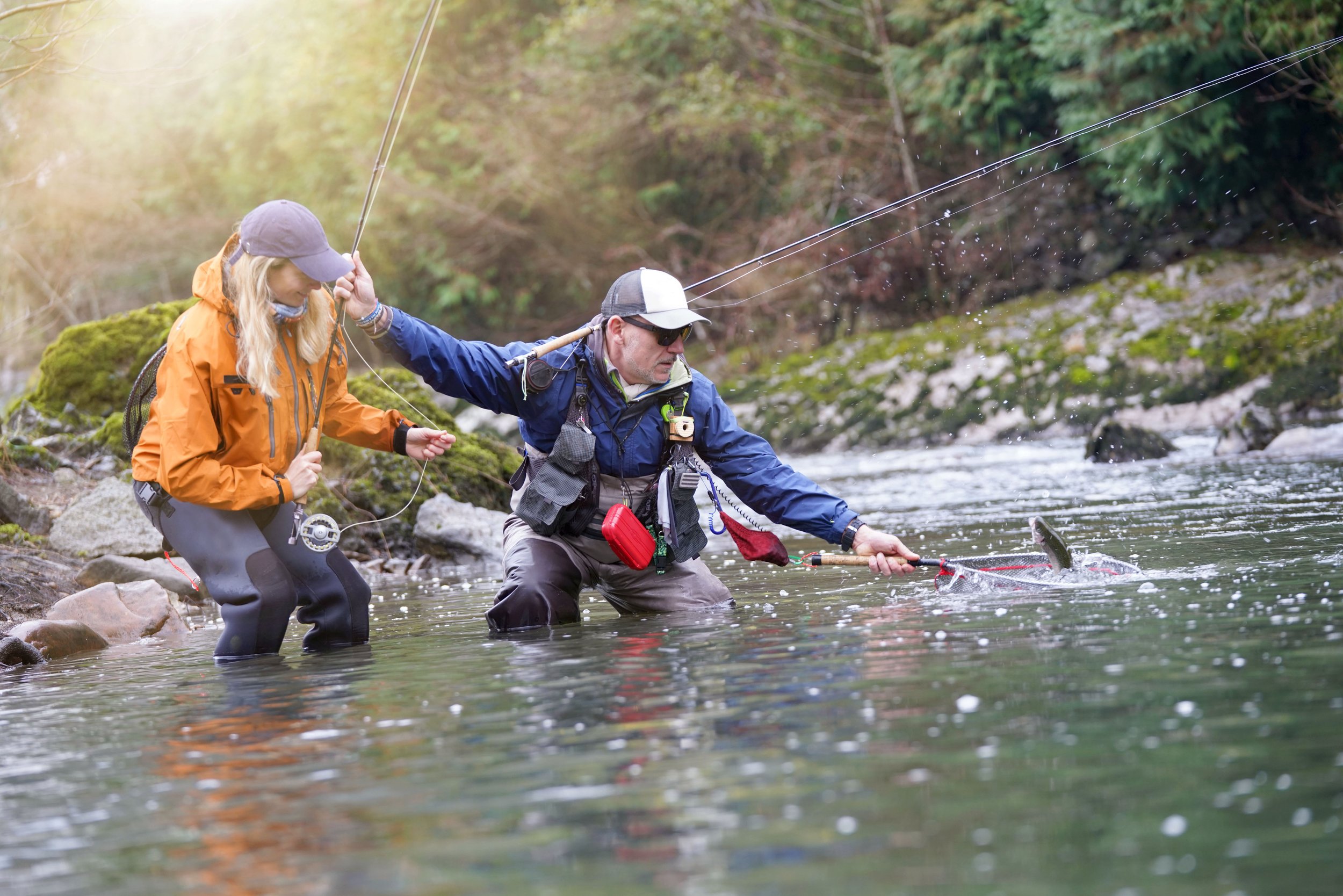 Two people fly fishing in a river, wearing outdoor fishing gear, one is netting a fish.