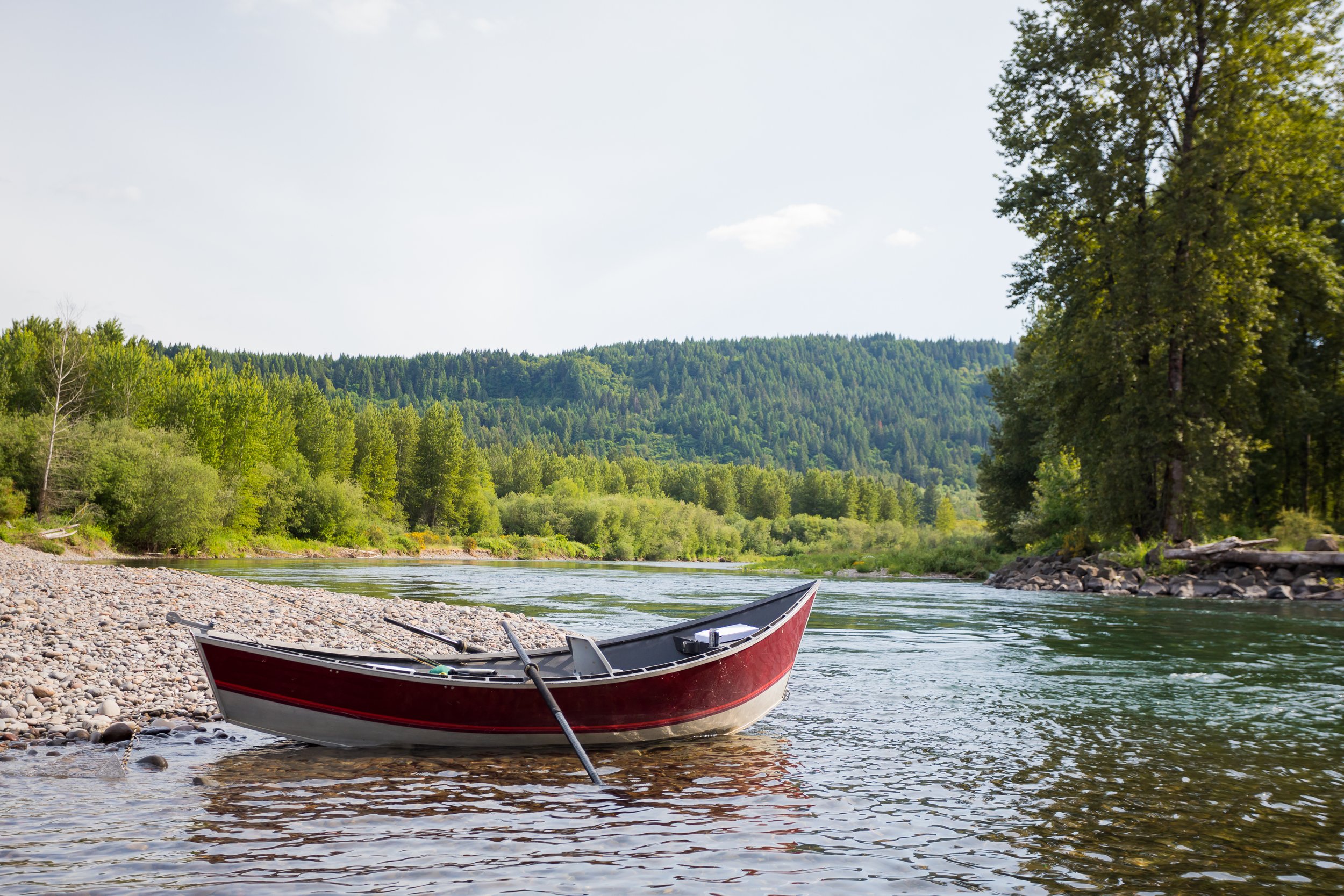 Red and white rowboat on a pebbled riverbank with forested hills and trees in the background.