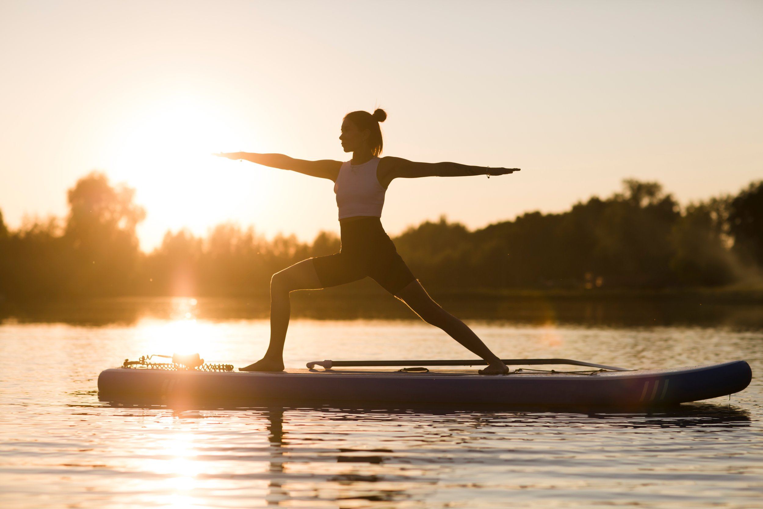 Silhouette of a person practicing yoga on a paddleboard at sunset on a lake, with trees in the background.