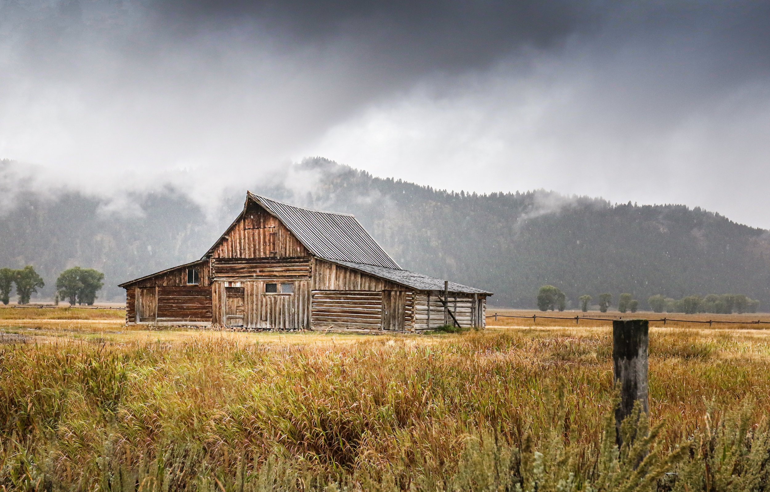 Old wooden barn in a field with mountains and cloudy sky in the background.