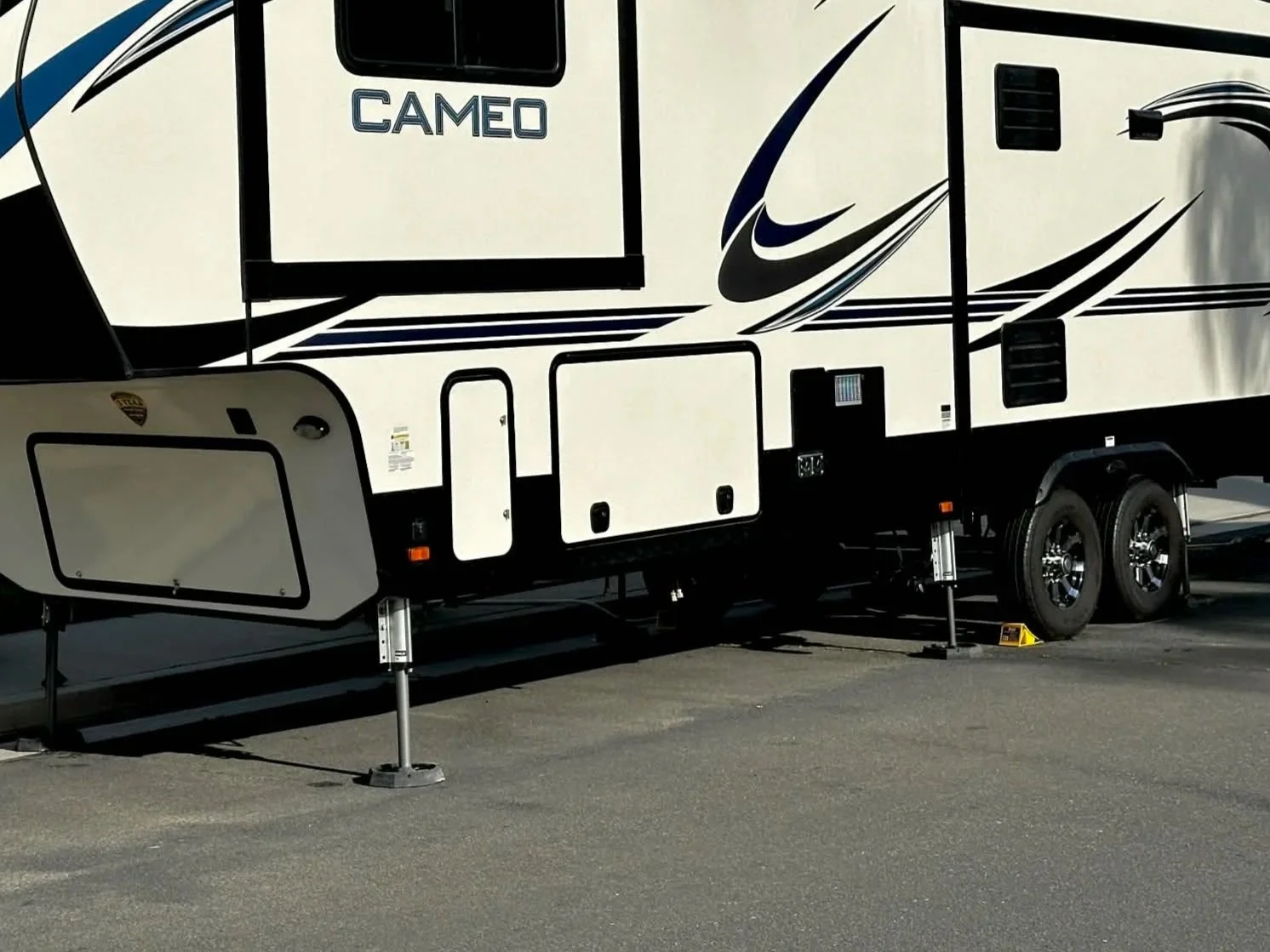 A large white and black travel trailer with the word 'CAMEO' on the side, parked on a paved lot with stabilizers underneath.