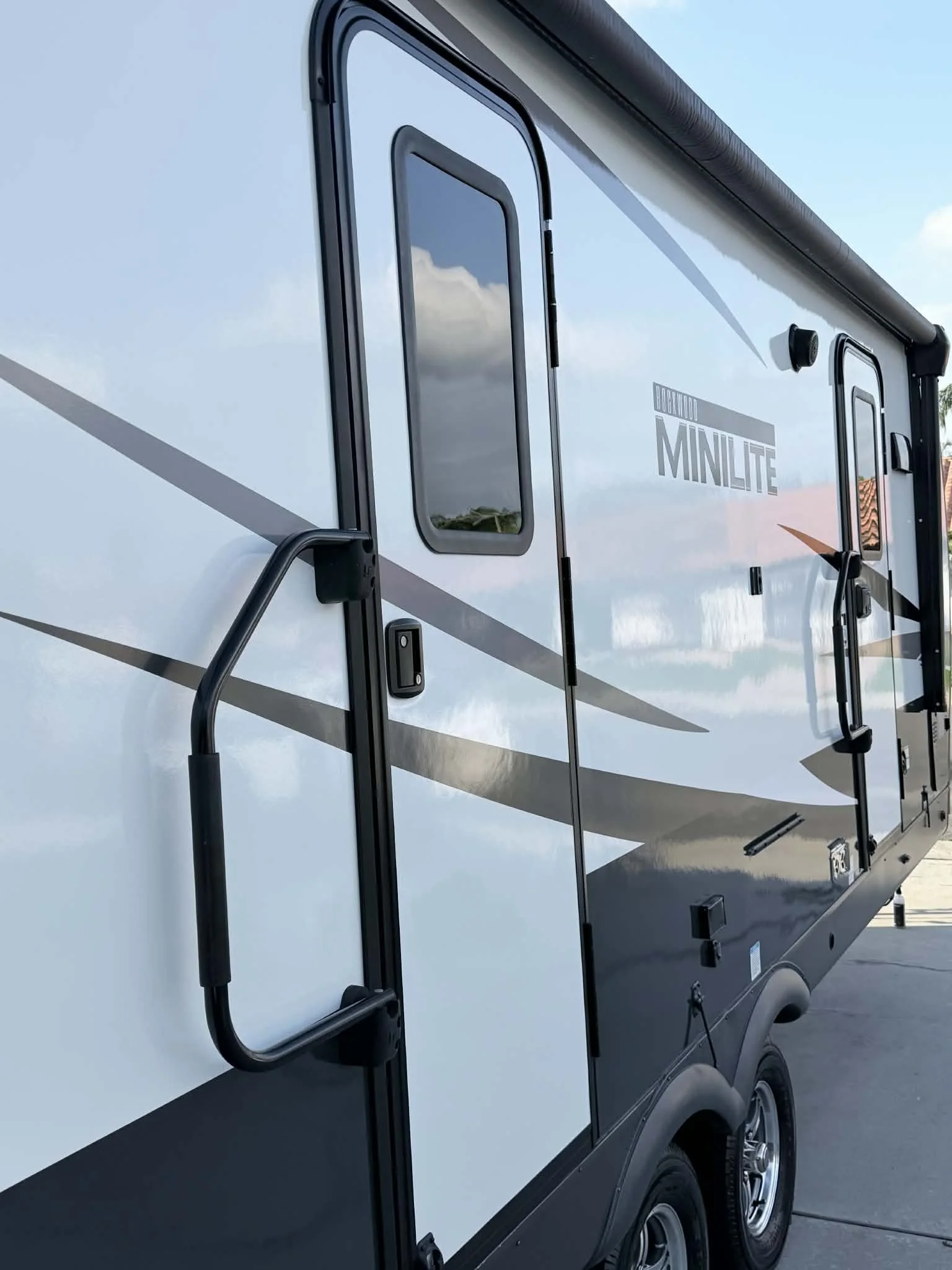 Close-up of a modern travel trailer with a black and white exterior, windows, door, and hardware, parked on pavement under partly cloudy sky.