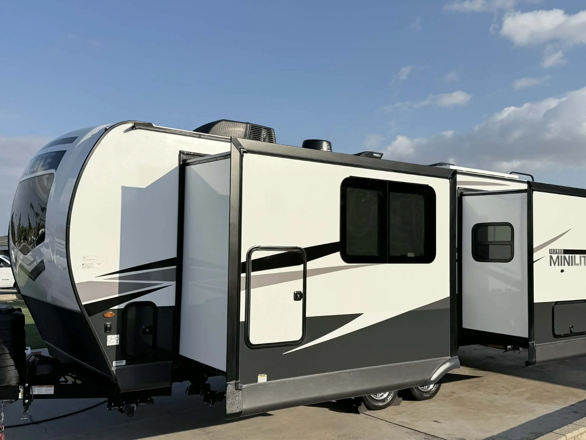 A modern travel trailer with white and black exterior parked outdoors on a paved surface, under a partly cloudy sky.