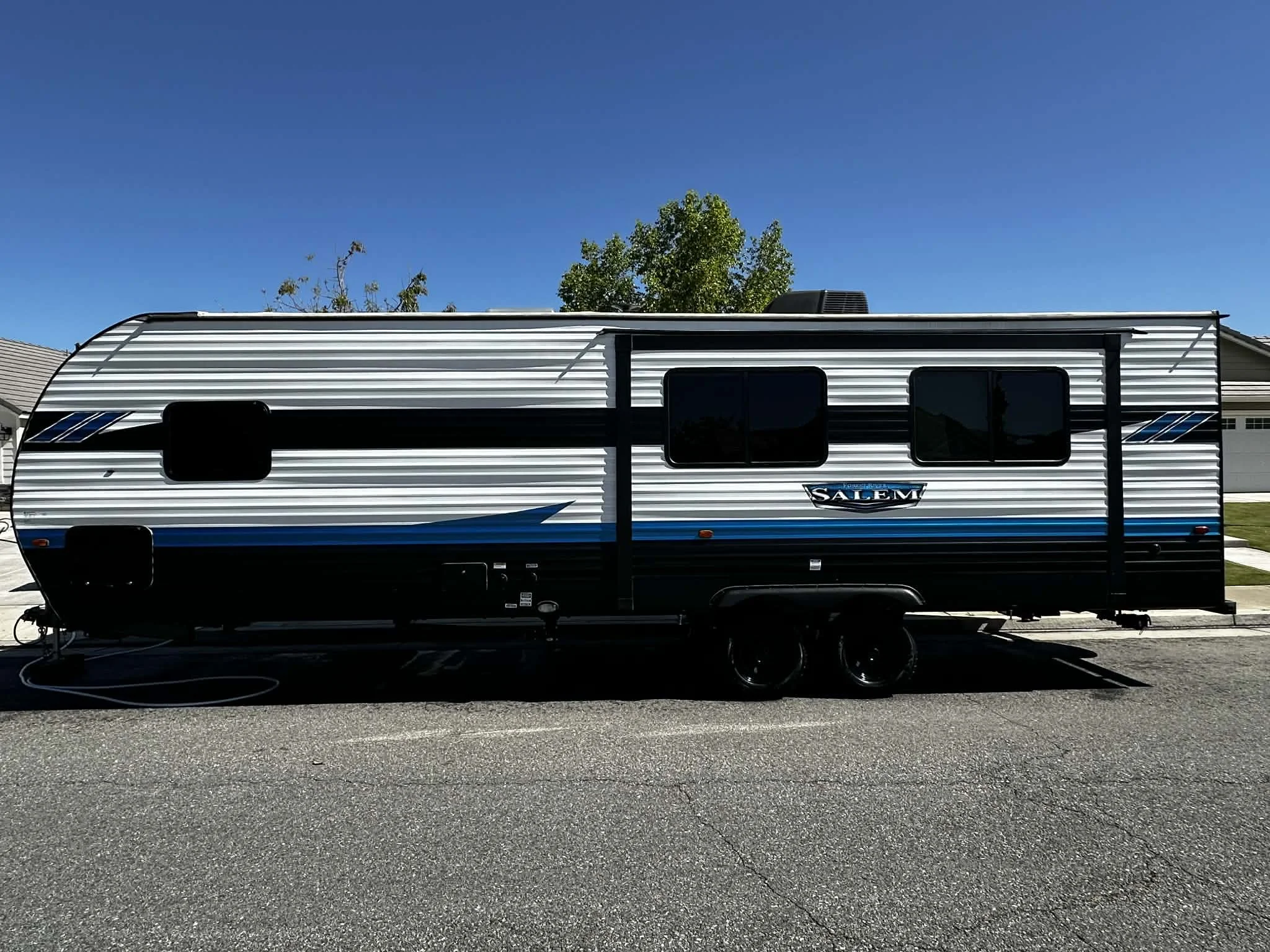 A black, white, and blue Salem travel trailer parked on a residential street with clear blue skies and a tree in the background.
