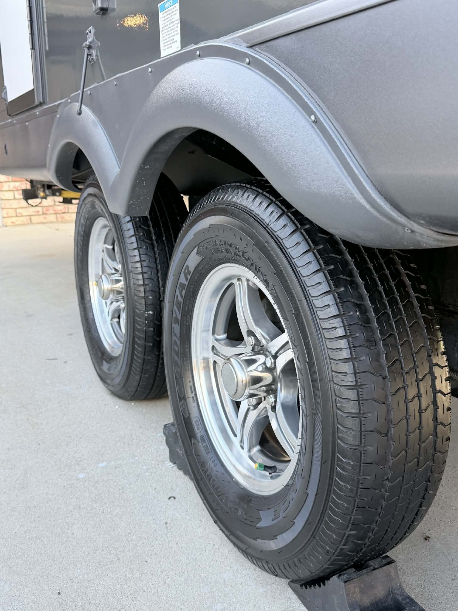 Close-up of a trailer's dual wheels, tires, and fenders, with a gray trailer body and brick wall in the background.