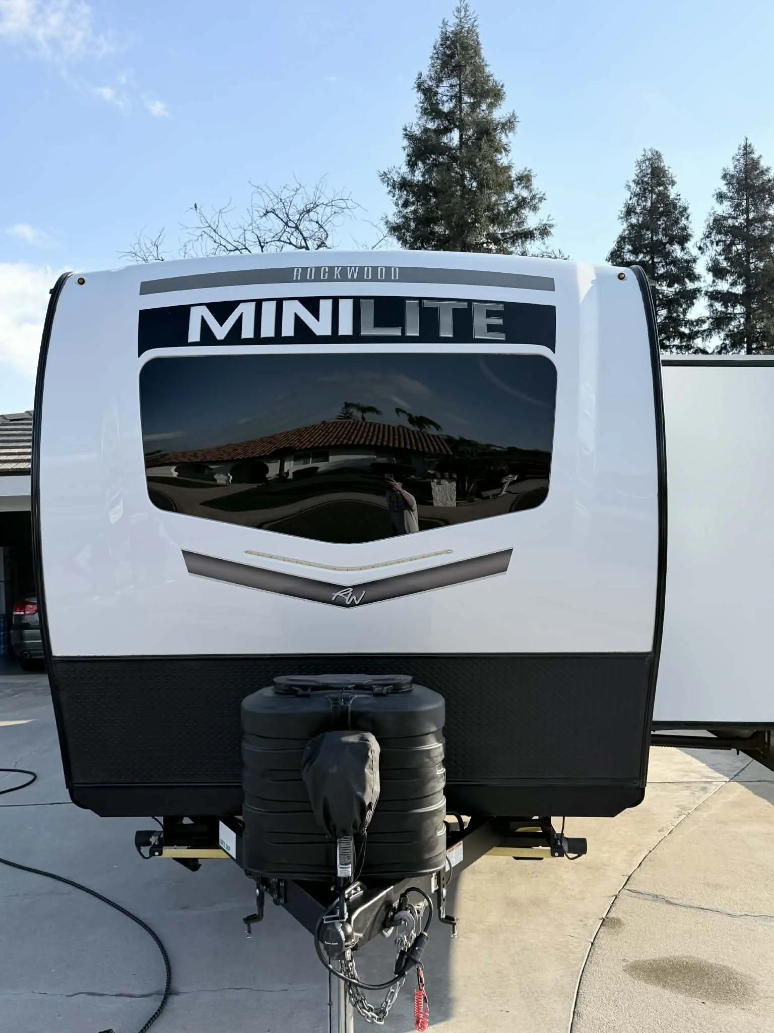 Front view of a Rockwood MiniLite travel trailer with a black rearview mirror reflected in the window, parked on a concrete surface with trees and a building in the background.