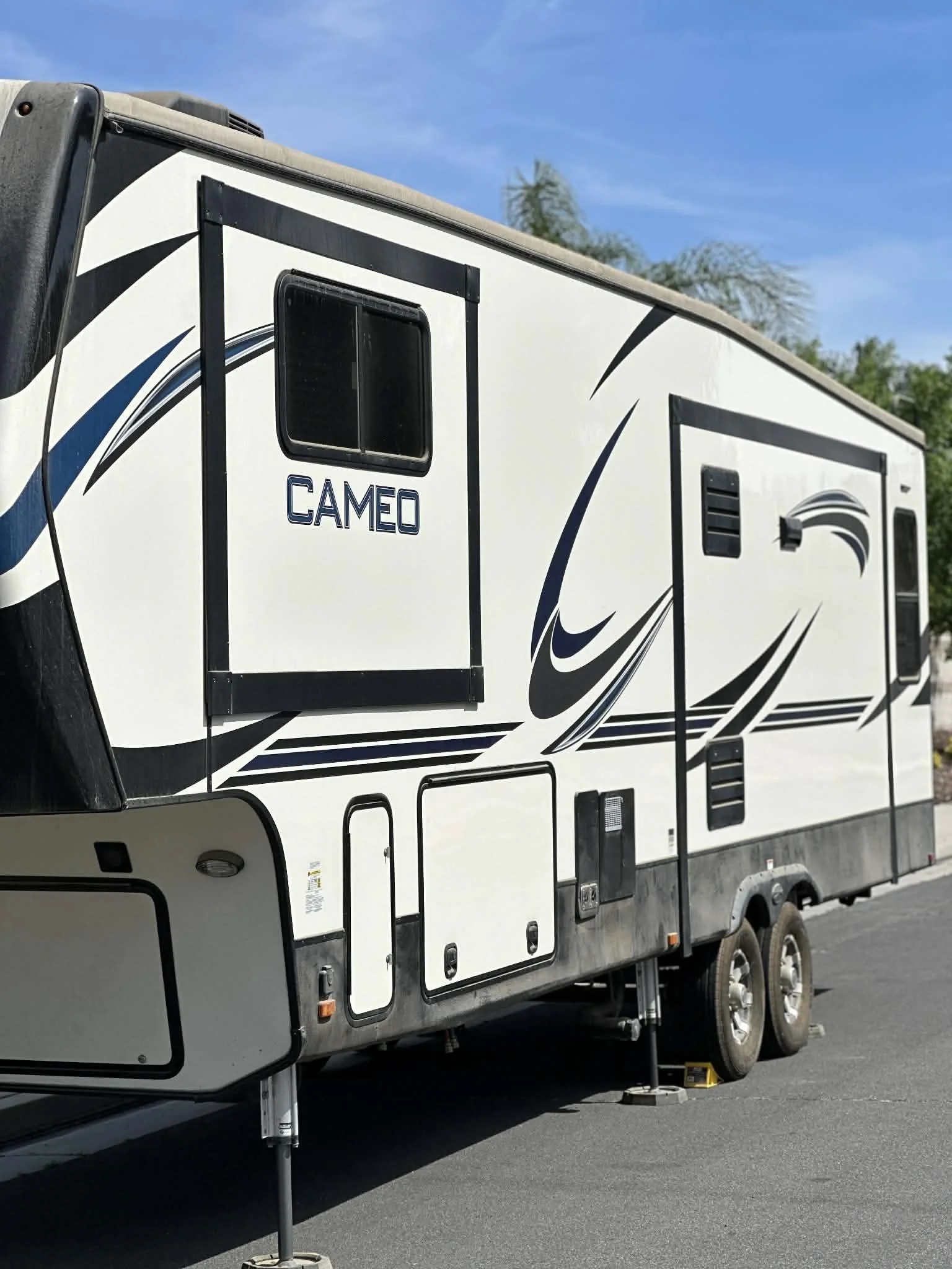 A white Cameo RV trailer with black and blue graphic accents parked on a paved surface under a blue sky with some trees in the background.