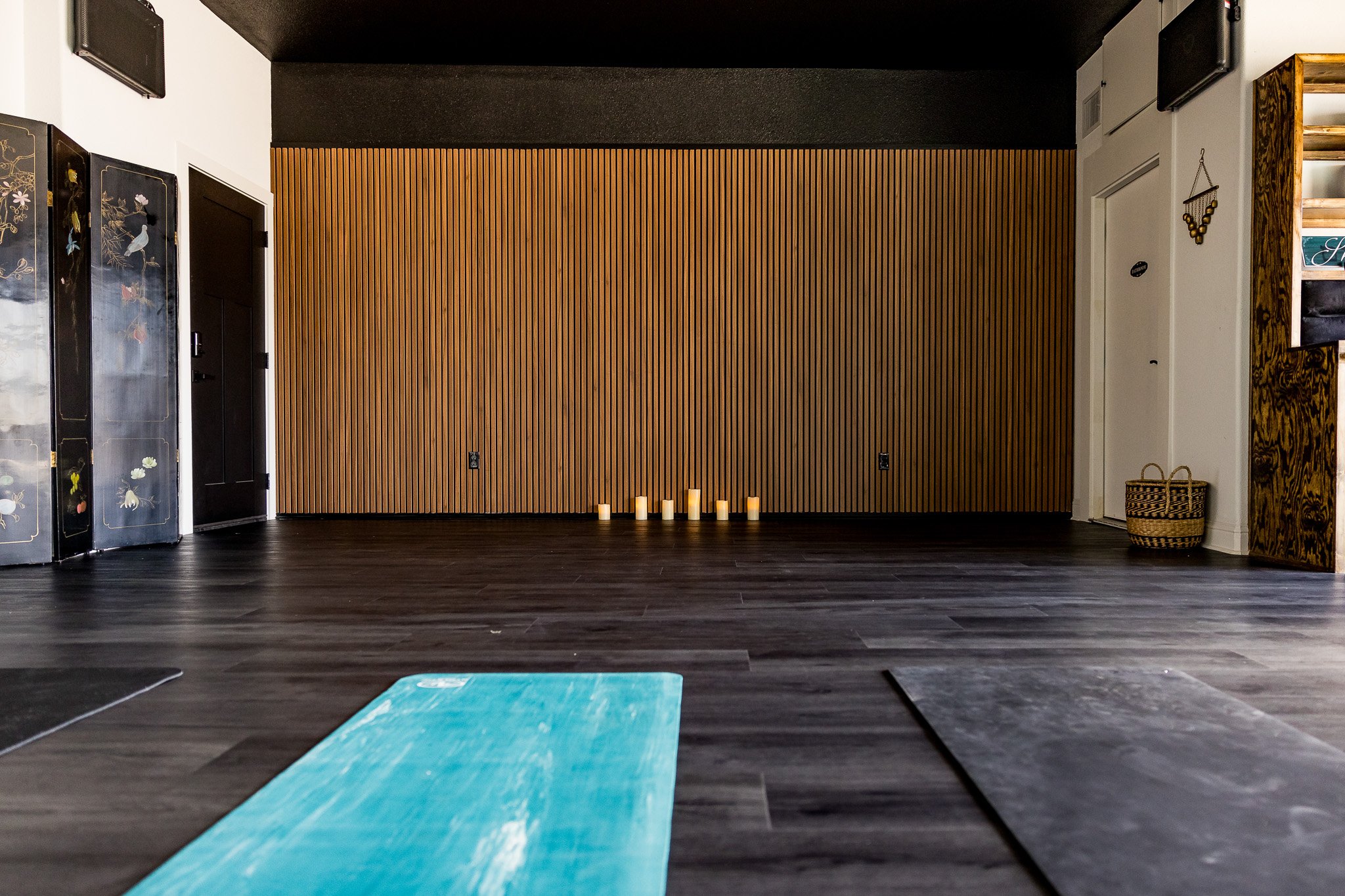 Empty yoga or meditation studio with wooden wall, incense candles on floor, yoga mats on dark wood floor, and decor on the walls.