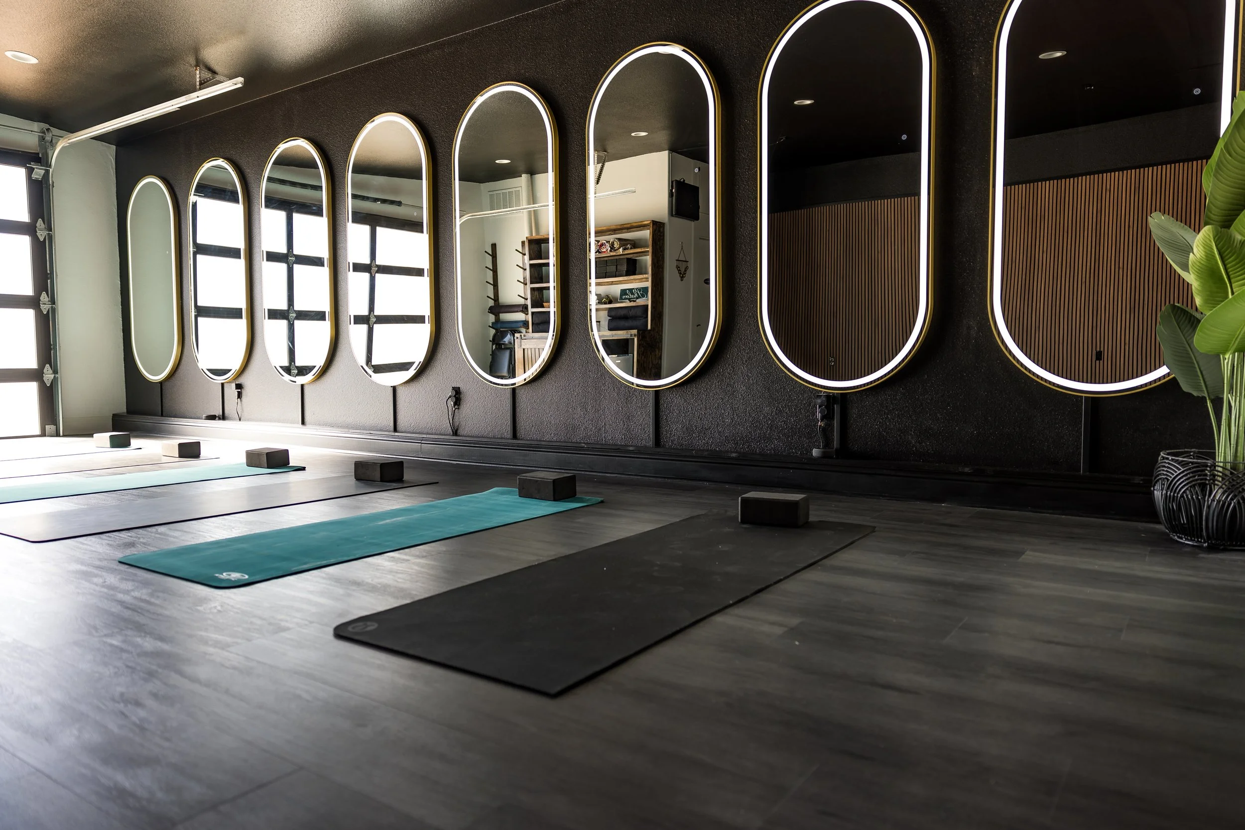 Yoga mats and blocks arranged in a studio with sleek oval mirrors and a dark wall, lit by natural light, with a potted plant in the corner.