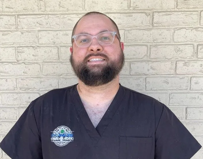 Man wearing glasses and a dark shirt with chiropractic logo, standing against a textured brick wall.