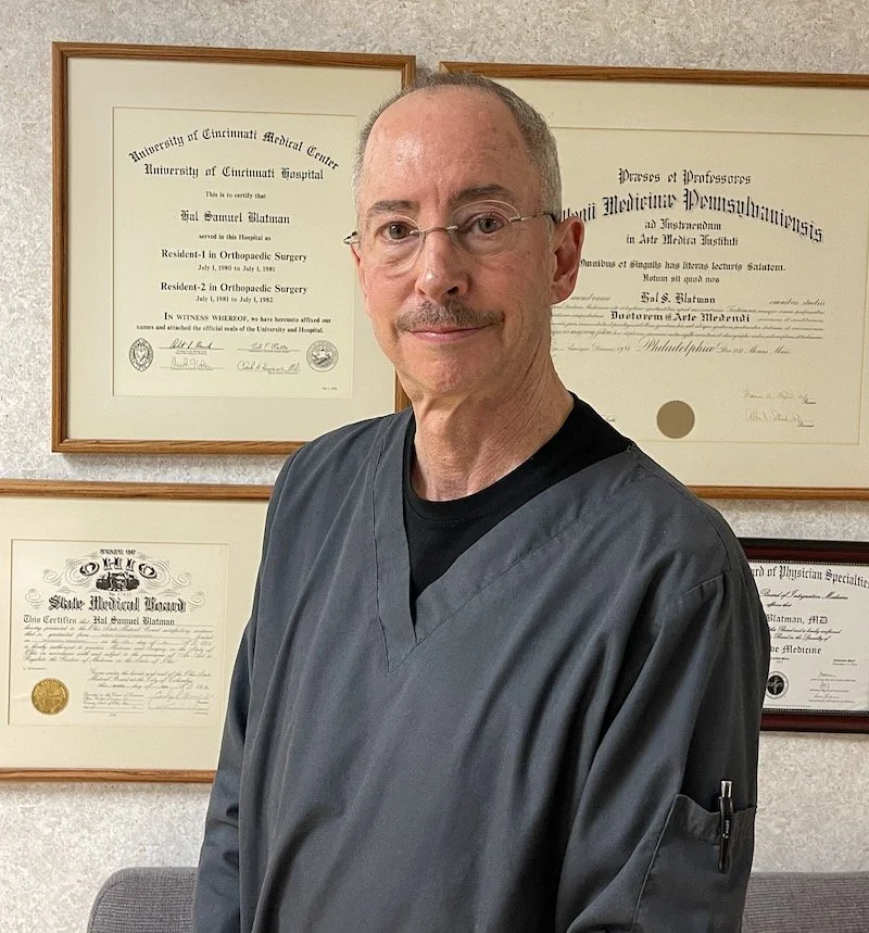 A person wearing gray scrubs stands in front of a wall with framed certificates.