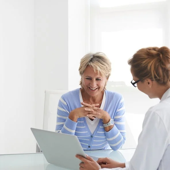 A woman in a blue striped sweater sitting at a desk, smiling, while talking to a woman in a white coat who is using a tablet.