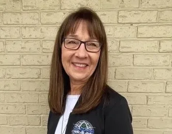 Smiling woman with glasses, long brown hair, wearing a black jacket, standing in front of a light-colored brick wall.
