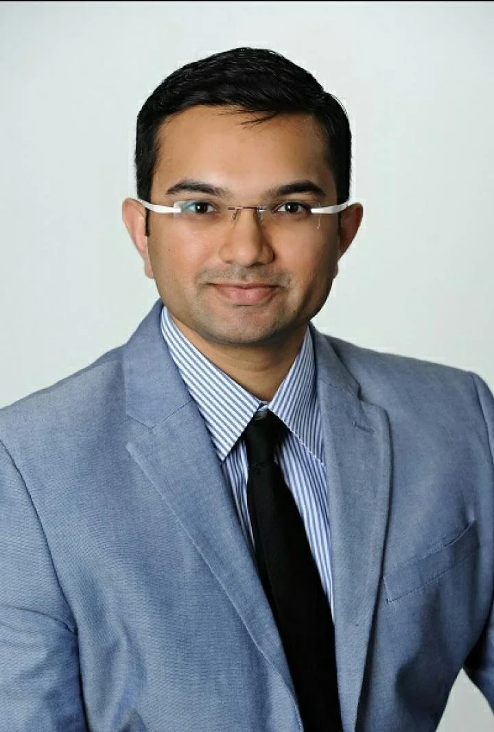 Portrait of a man wearing glasses, a light blue suit, white and blue striped shirt, and black tie.