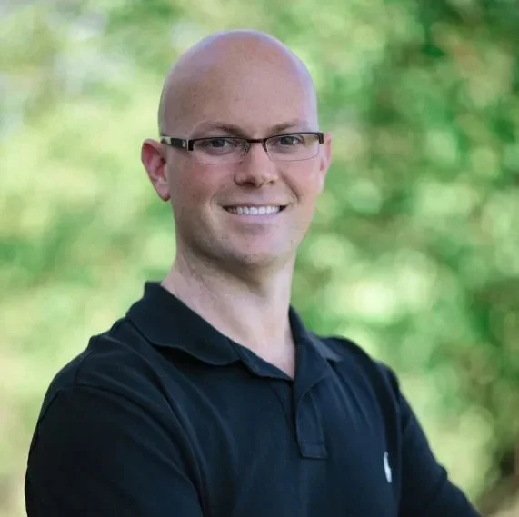 Bald man with glasses in black shirt smiling outdoors.