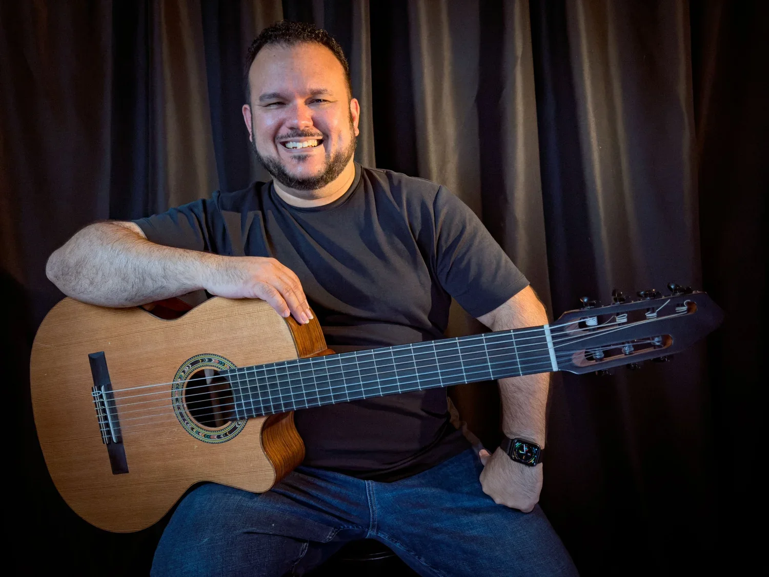 A smiling man in a black T-shirt holds a classical guitar against a dark curtain backdrop.