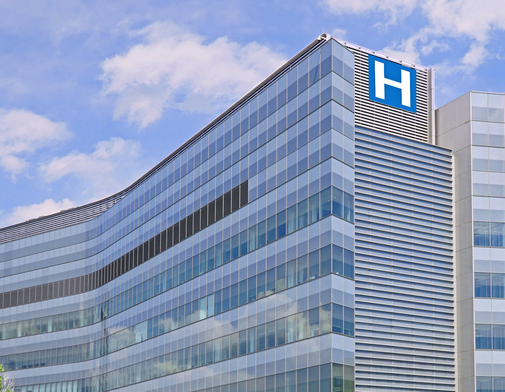 Modern glass hospital building with a blue H sign on the corner, set against a background of a partly cloudy sky.